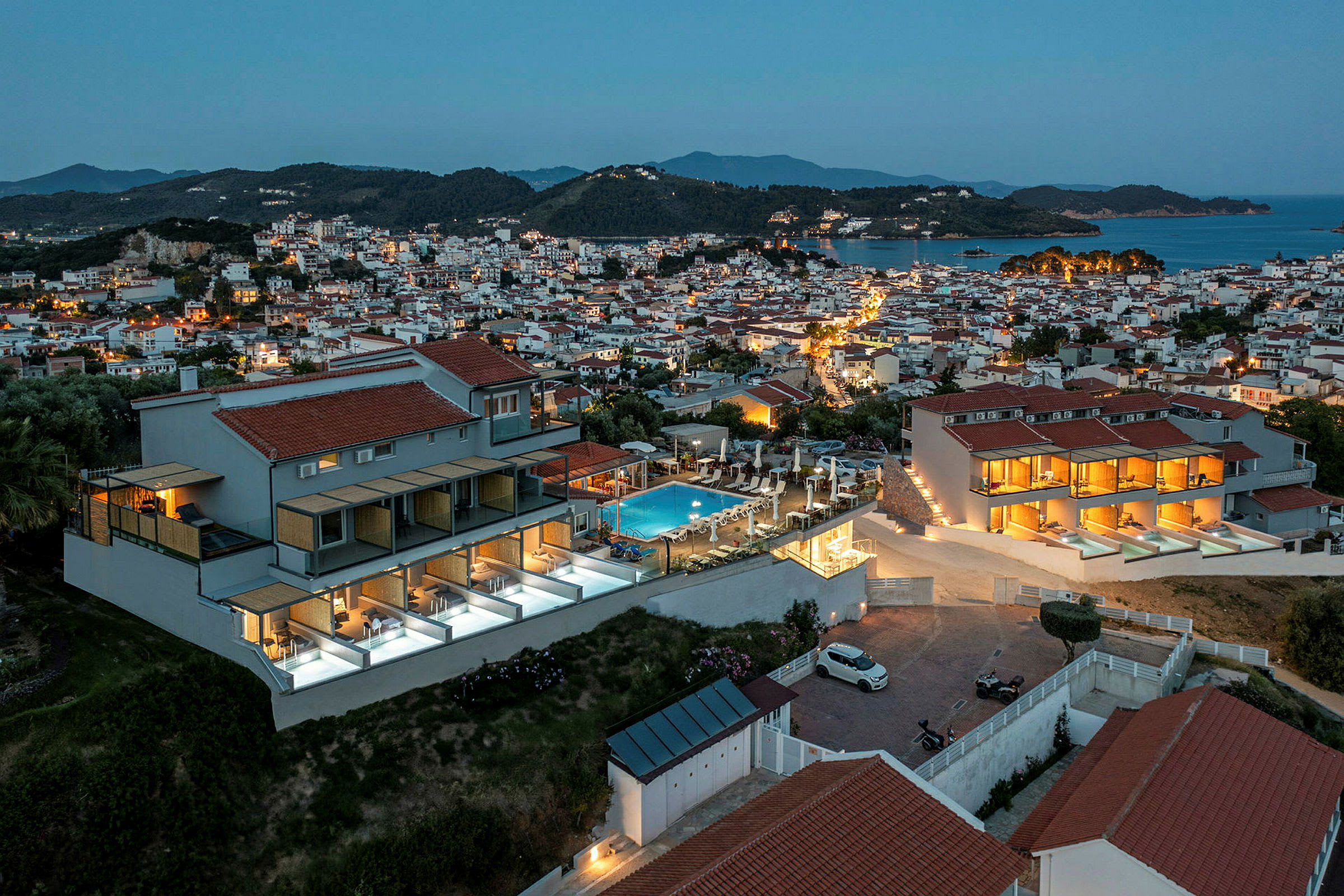 Bild föreställer en vy över en stad vid skymning, med hus med rödaktiga tak, kullar i bakgrunden och en pool i förgrunden. (AI-genererad text)