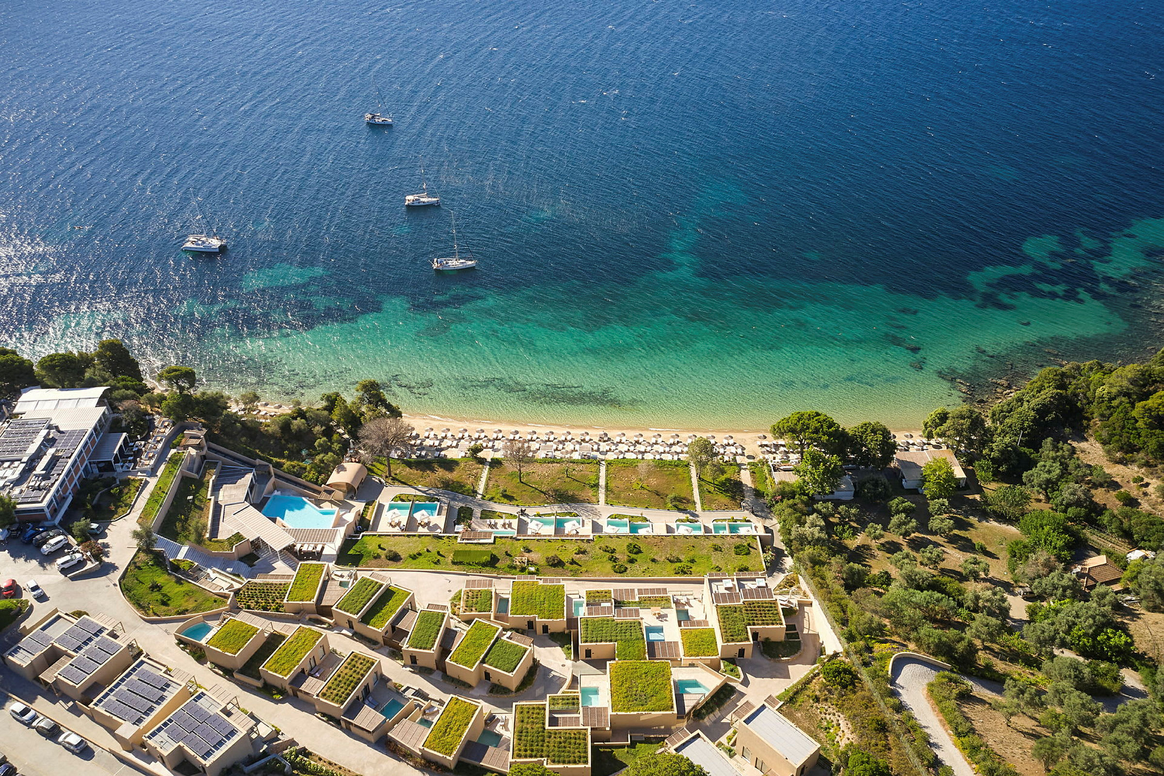 Vy från ovan av en kustlinje med blått hav, strand med solstolar samt semesterbyggnader med grönskande tak. (AI-genererad text)