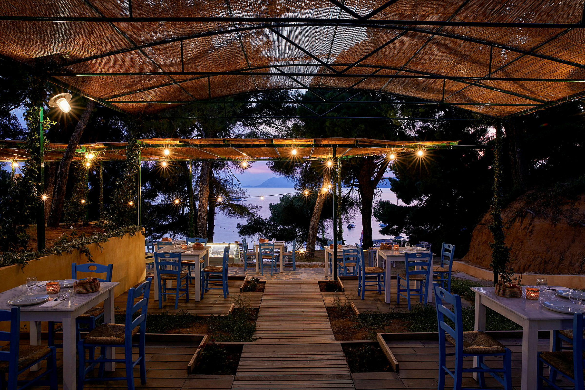 Utomhusrestaurang med blå stolar och vita bord, belägen nära havet på kvällen. (AI-genererad text)