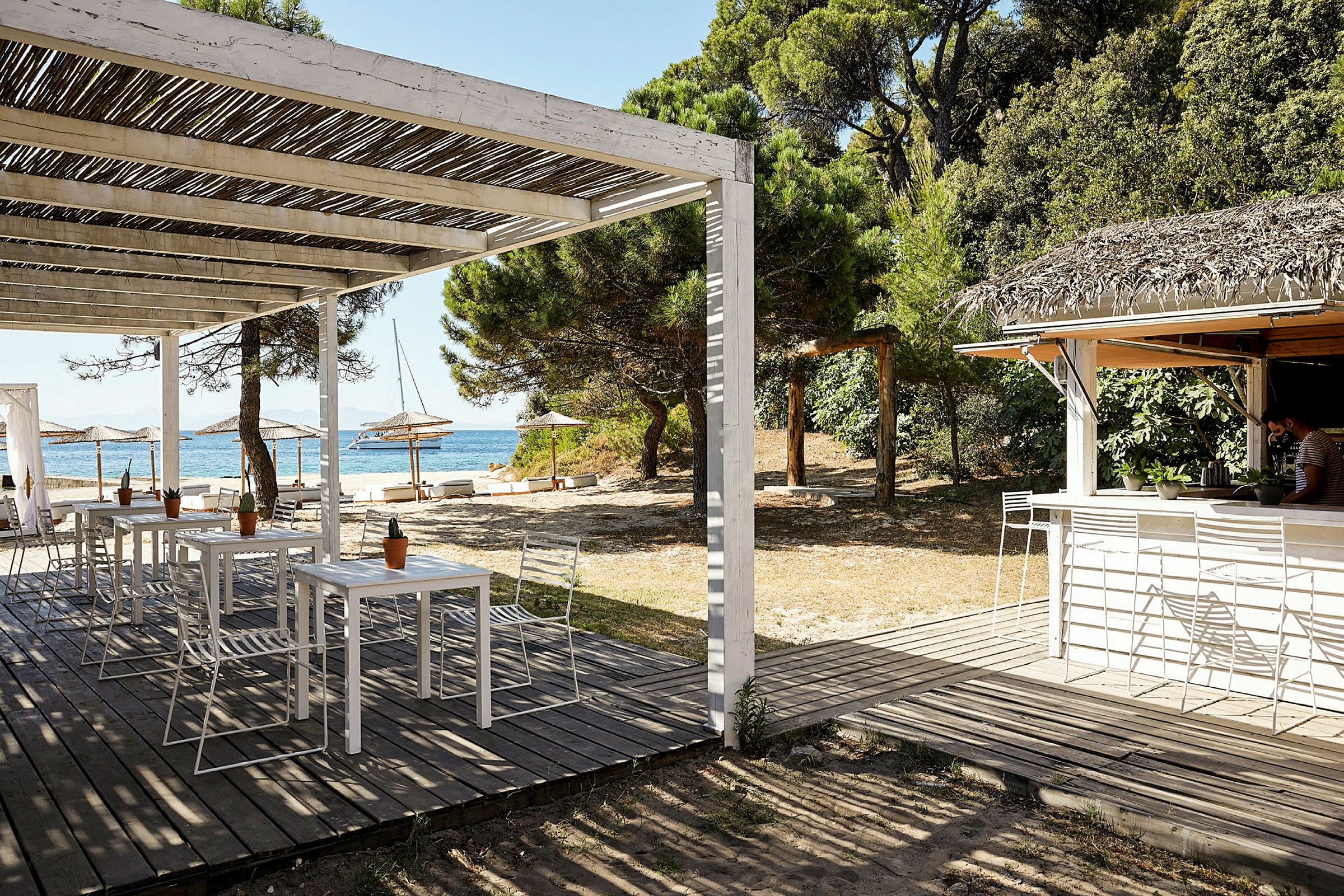 En strandbar med sittplatser under en träpergola, närliggande parasoller och en skog i bakgrunden. (AI-genererad text)