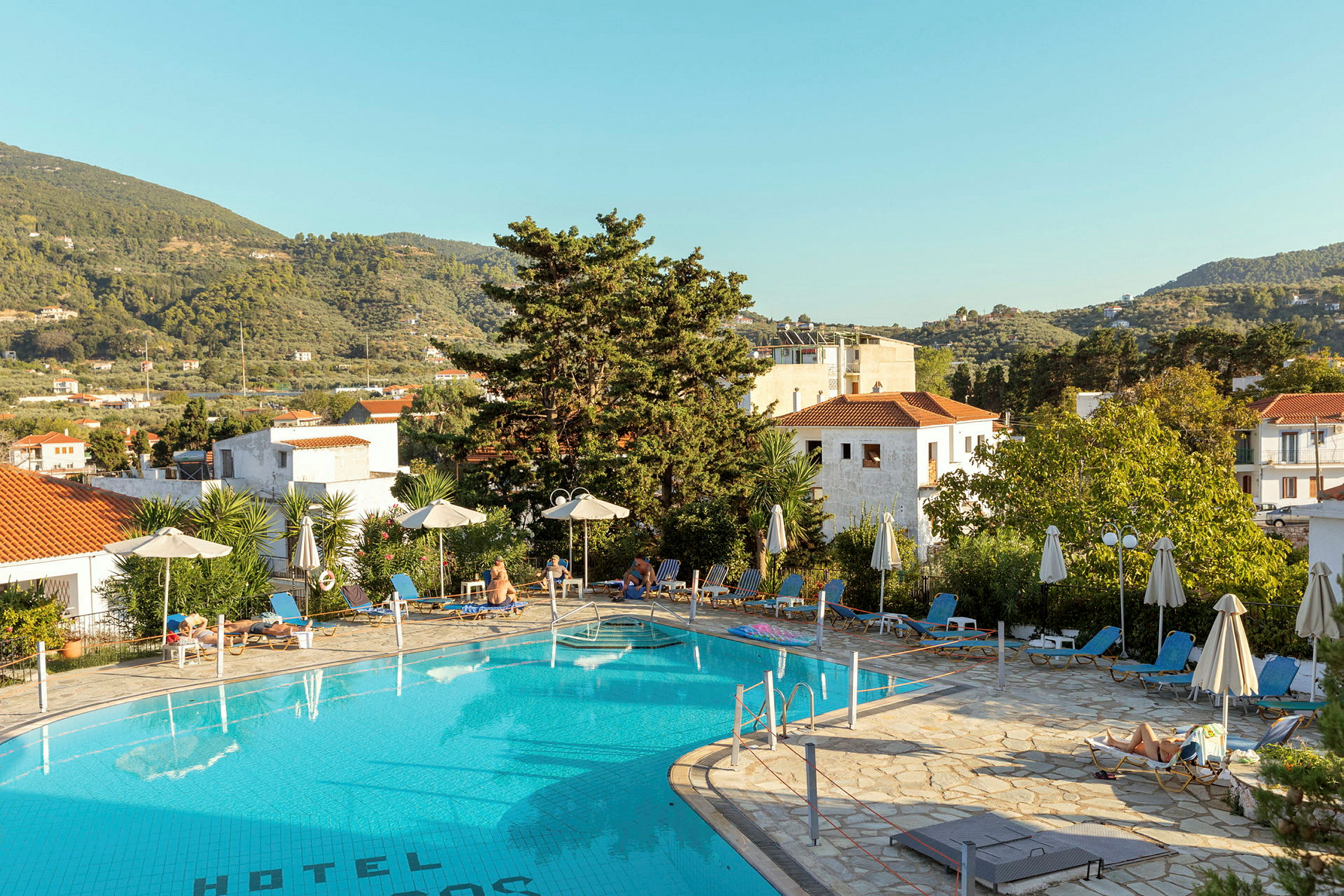 Utomhuspool vid hotell med solstolar och parasoller, omgiven av grönska och berg. (AI-genererad text)