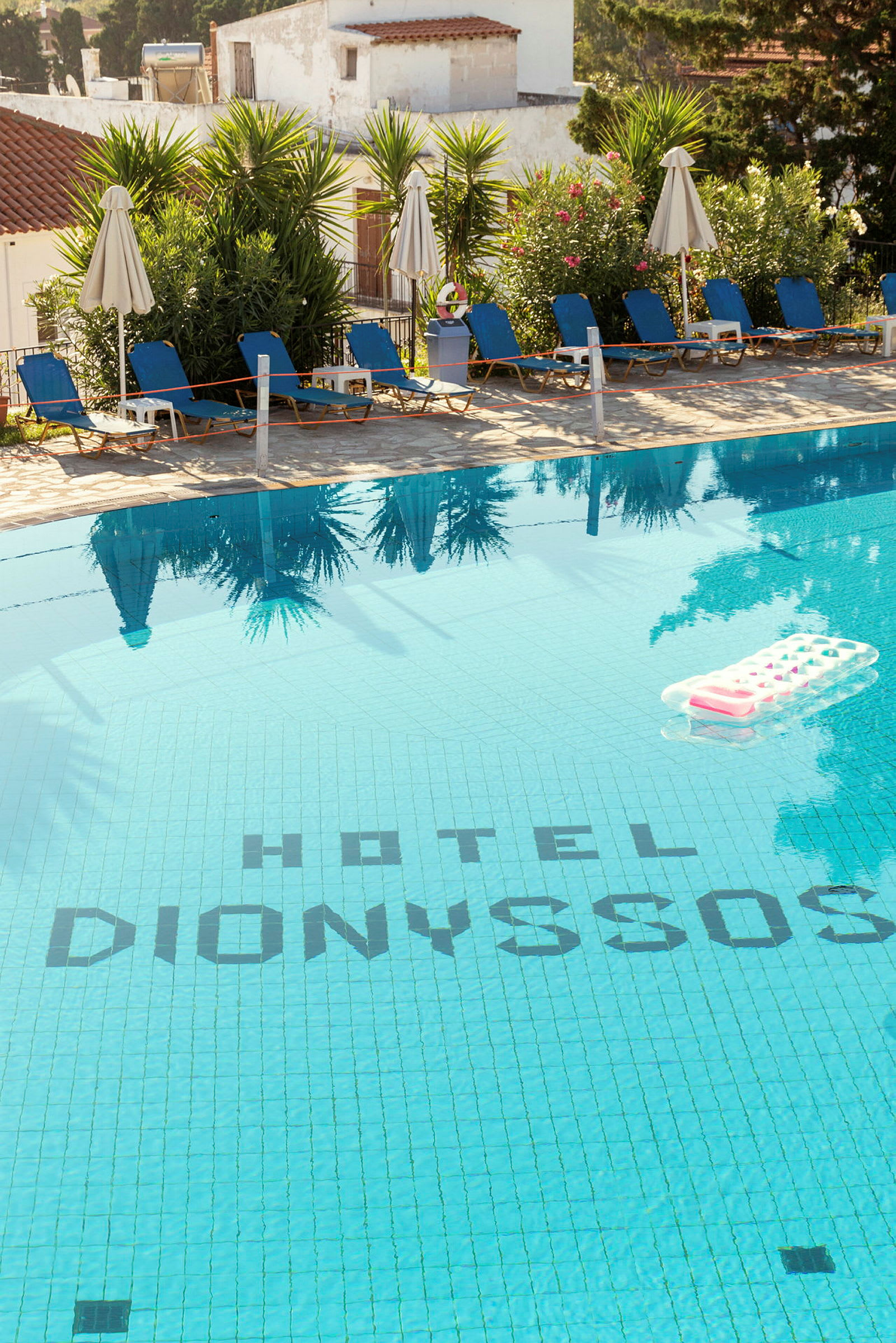 Utomhuspool vid Hotel Dionysos med solstolar och parasoller omgivna av grönska. (AI-genererad text)
