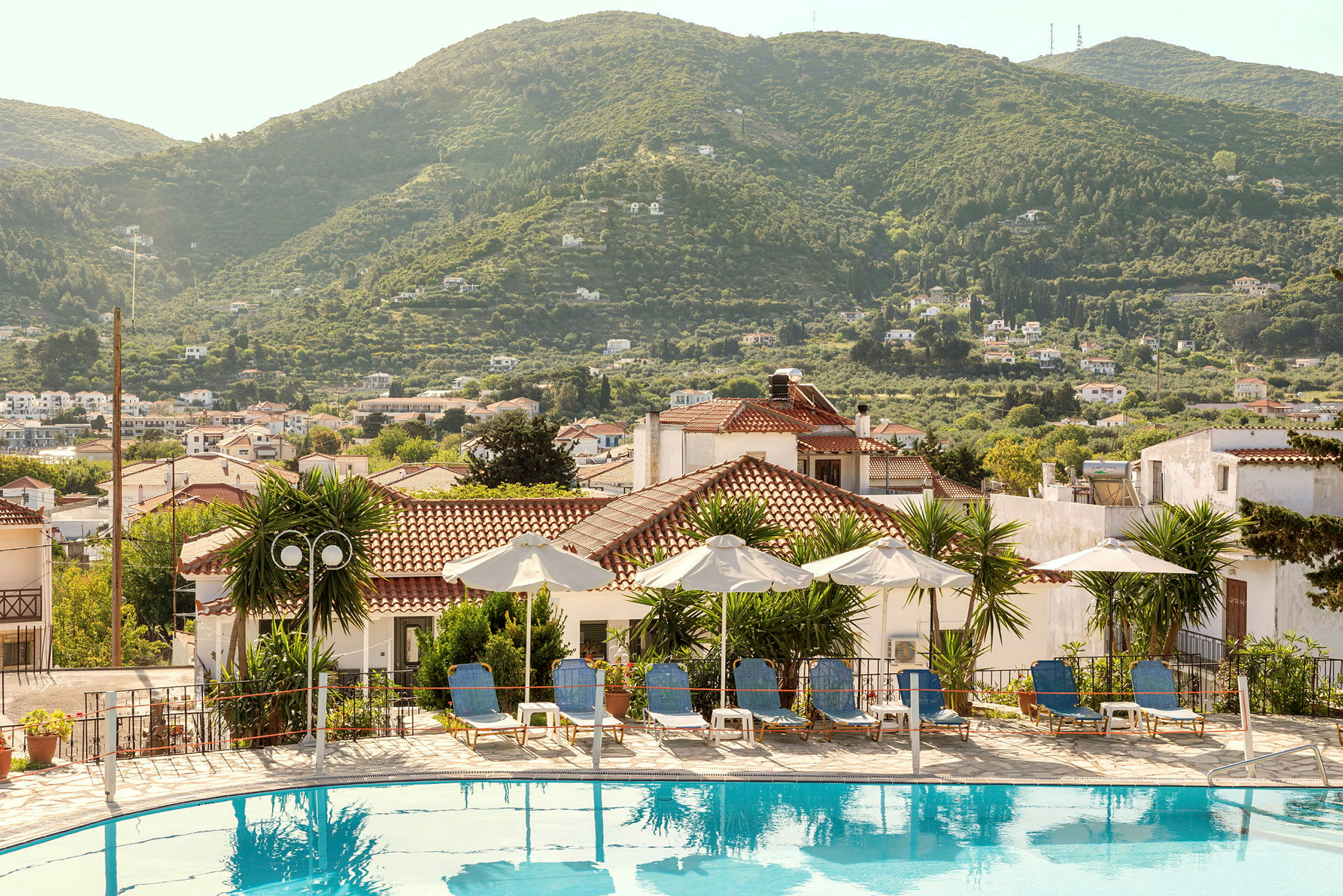 Pool med solsängar och parasoller, omgiven av hus och grönskande kullar. (AI-genererad text)