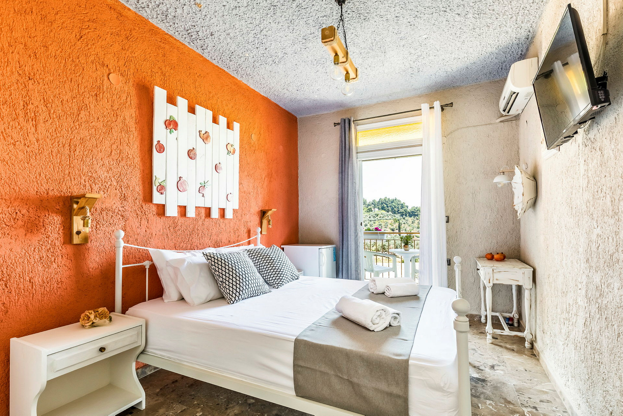 Hotellrum med en orange accentvägg, dekorerad med en vit träram med fruktmotiv, en säng med vita lakan och grå överkast, och en liten balkong med utsikt mot grönska. (AI-genererad text)