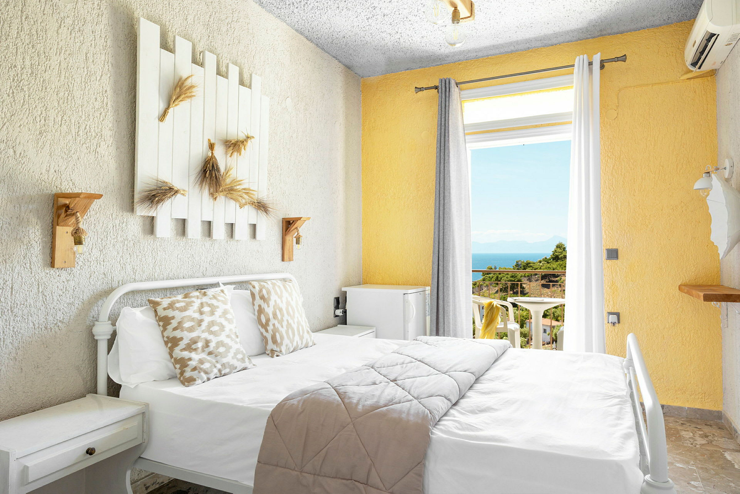 Ljust hotellrum med säng, dekorerade kuddar och utsikt mot balkong med hav och grönska. (AI-genererad text)