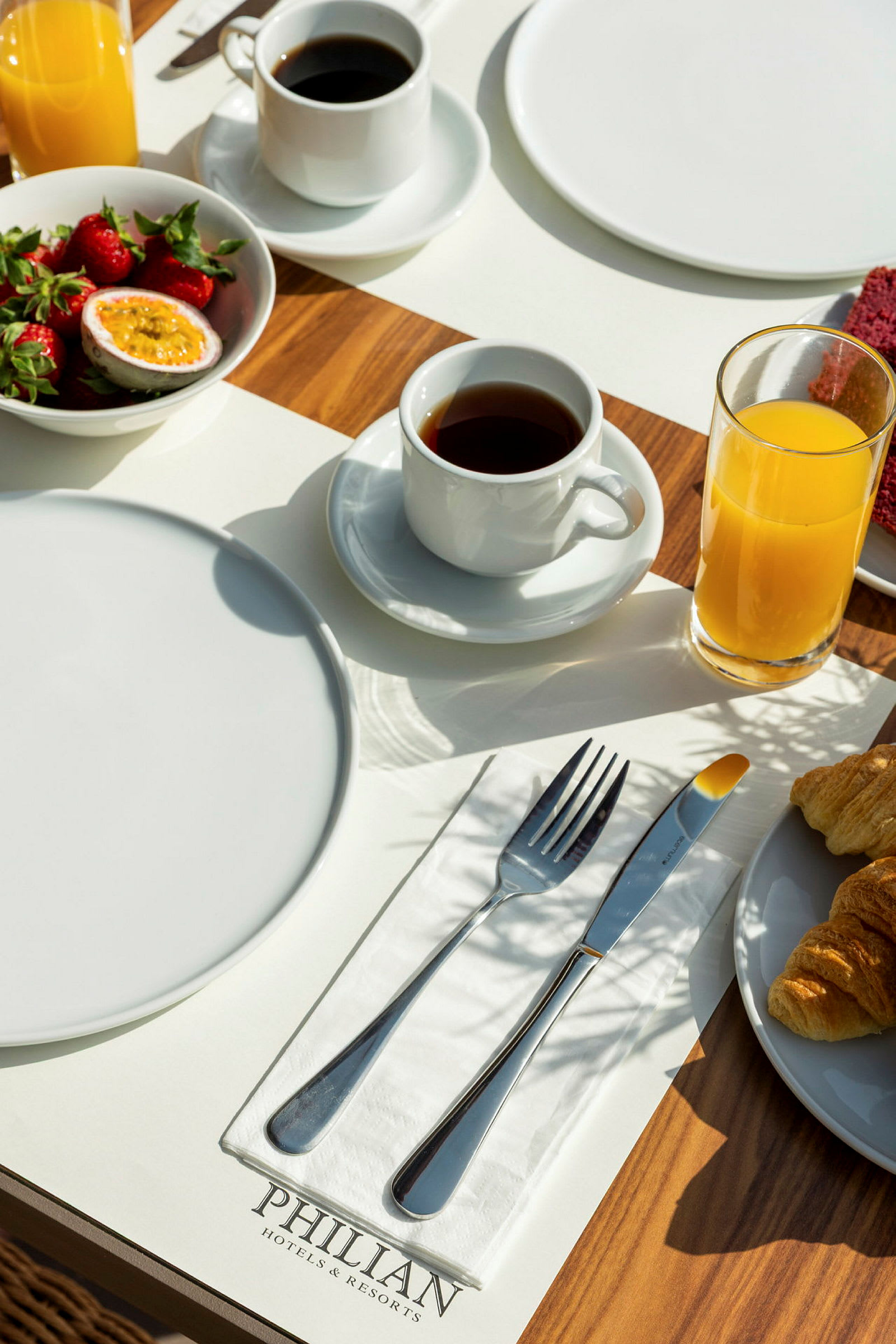 Frukostbord med tallrikar, bestick, kaffe, juice, jordgubbar och croissanter på en träyta. (AI-genererad text)