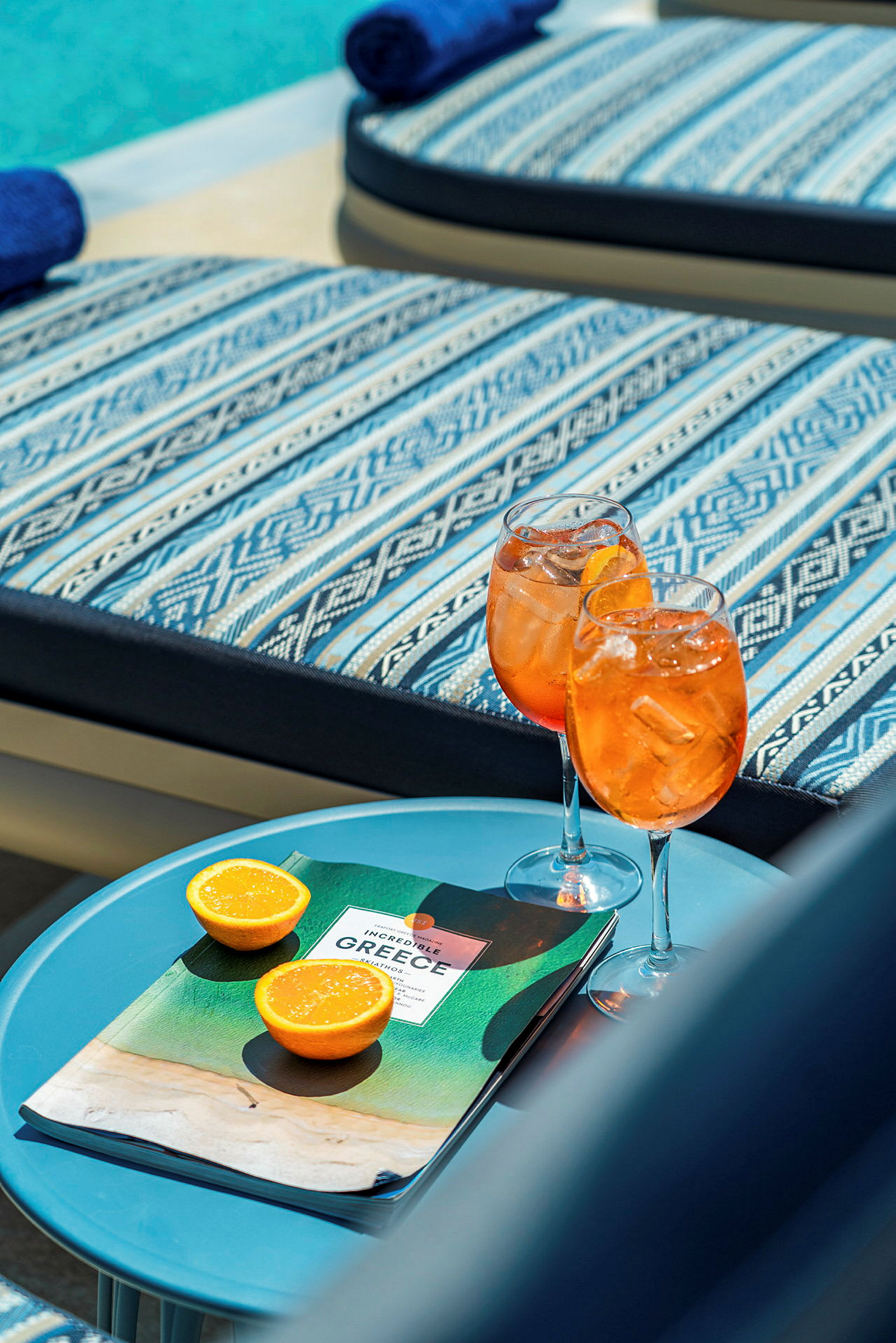 Två glas med orange dryck, en broschyr och två halvor apelsin på ett bord bredvid solstolar. (AI-genererad text)