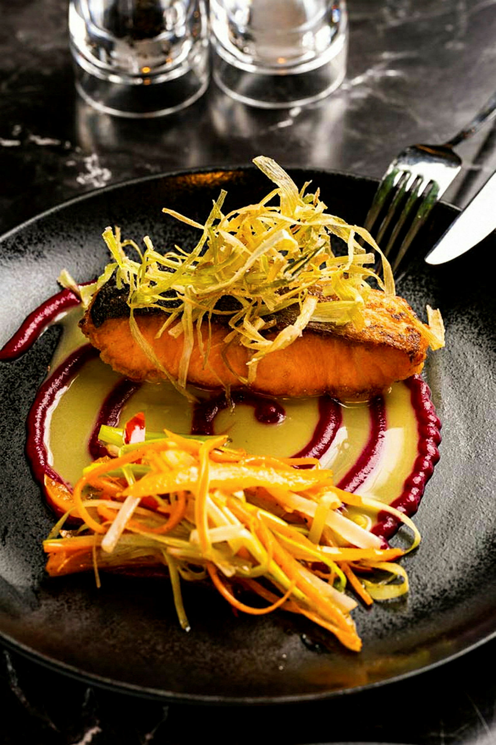 En tallrik med en maträtt bestående av tillagad fisk, topping av grönsaker och sås. Bestick och glas syns i bakgrunden. (AI-genererad text)