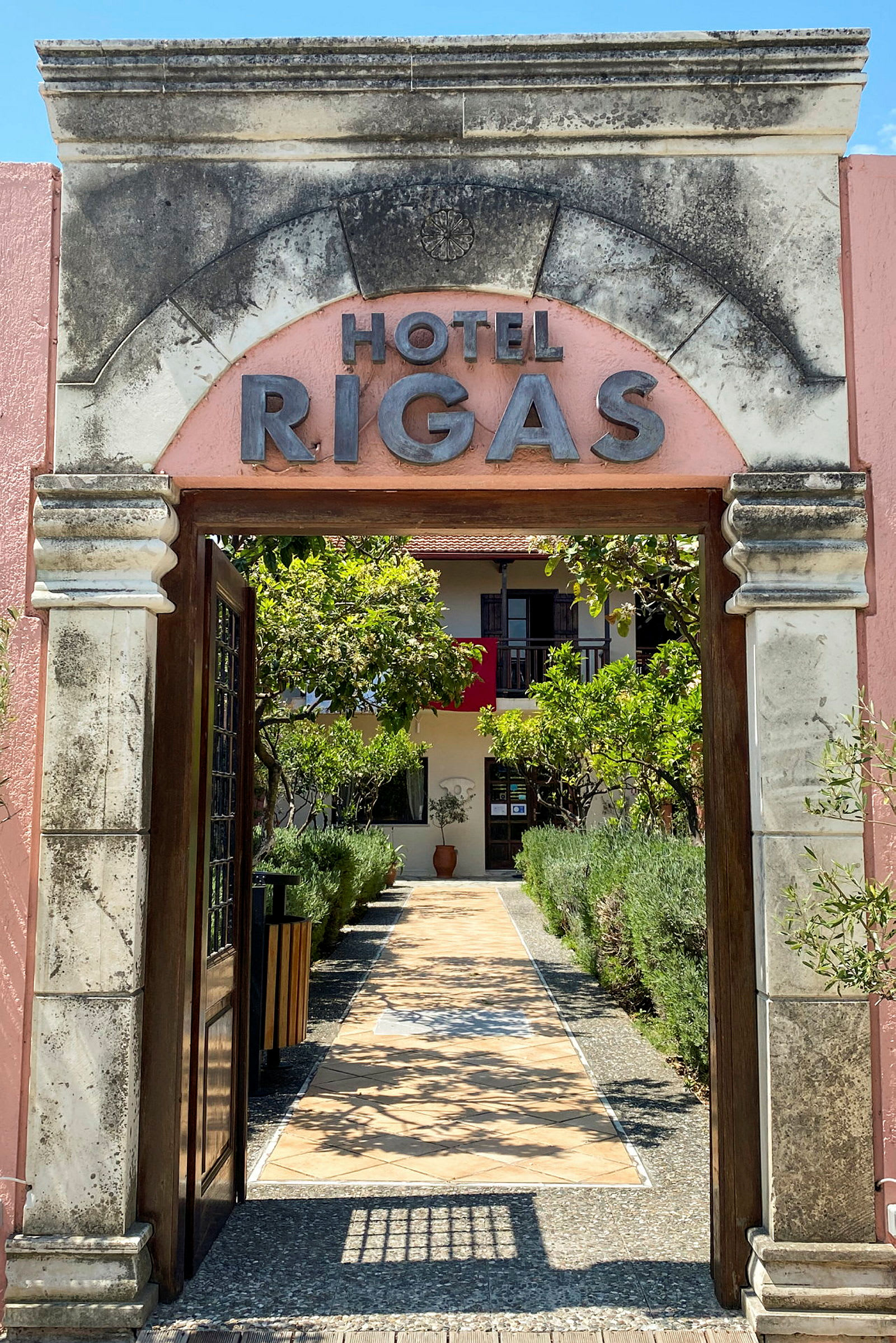 Bild av en hotelldörr med texten 'Hotel Rigas', omgiven av en trädgård och en stig mot ingången. (AI-genererad text)