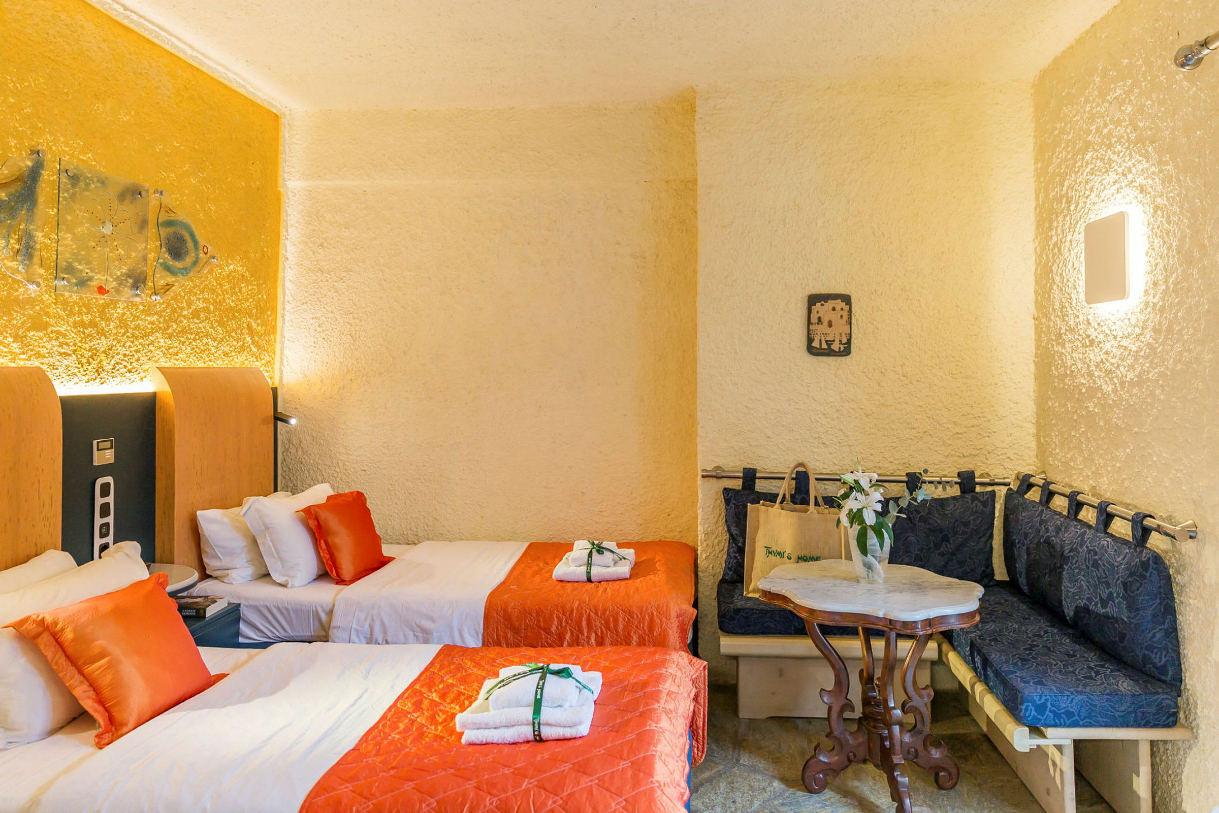 Ett hotellrum med två enkelsängar, orange sängöverkast och kuddar, samt en sittgrupp med ett bord och stolar. (AI-genererad text)