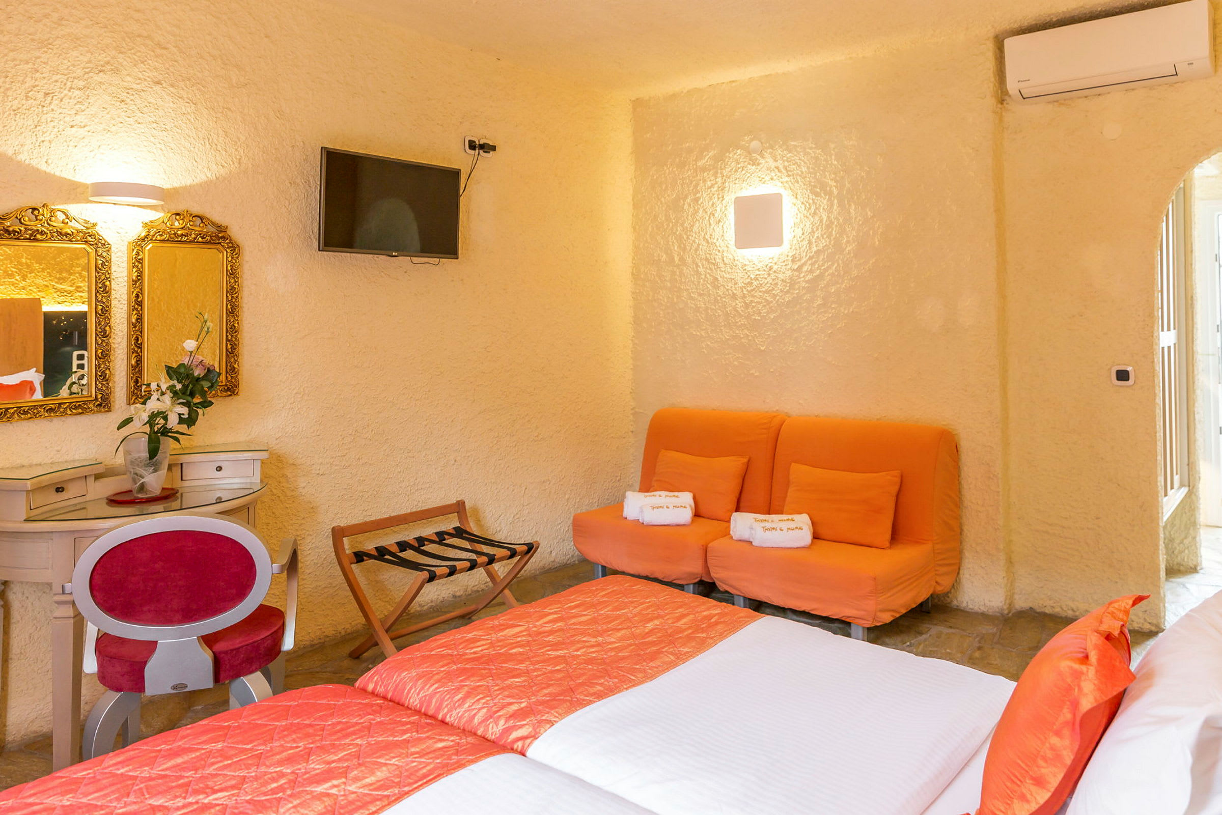Ett hotellrum med två sängar, en orange soffa, och ett sminkbord med speglar. (AI-genererad text)
