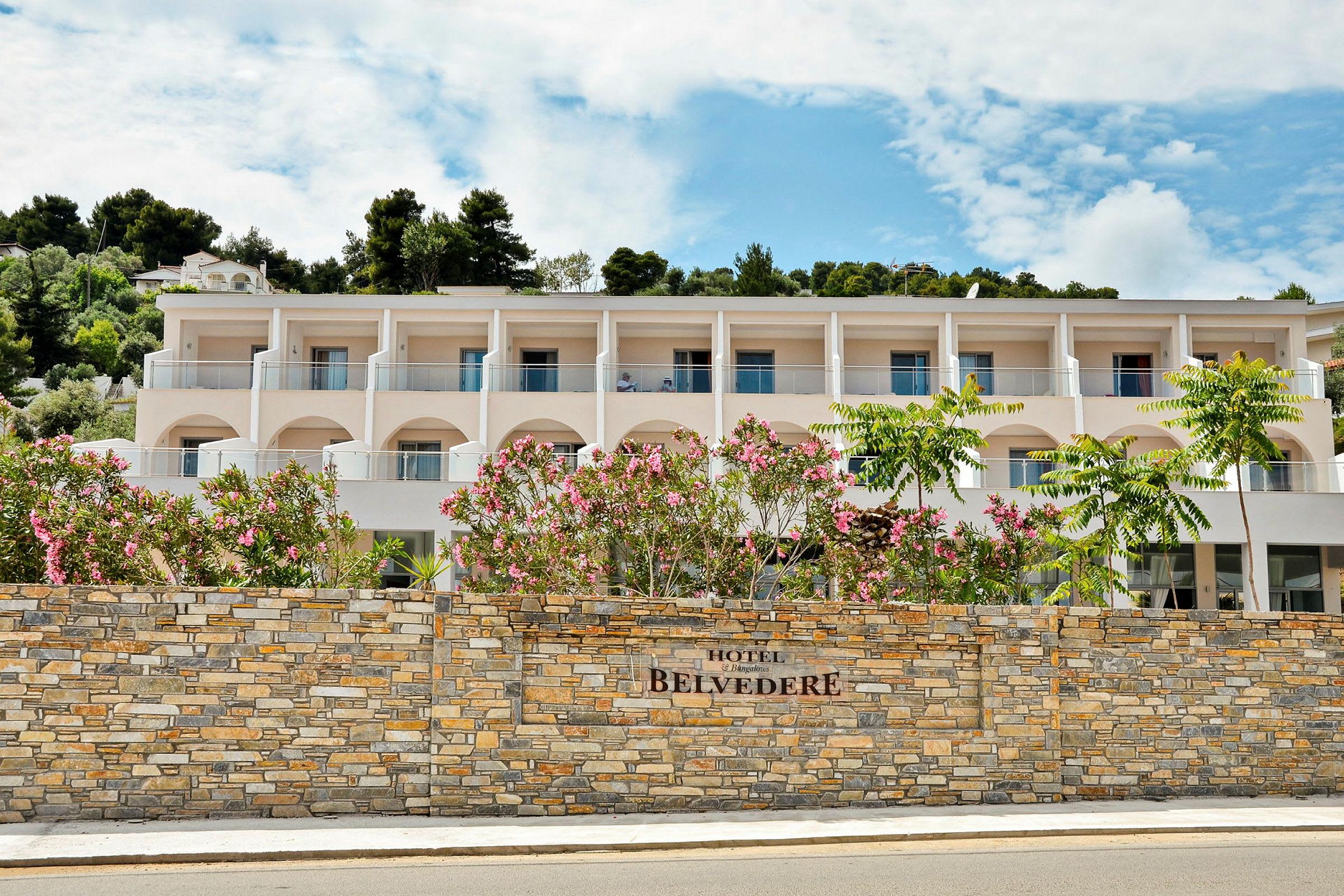 Hotellets fasad med balkonger och en mur framför samt blommande buskar. (AI-genererad text)