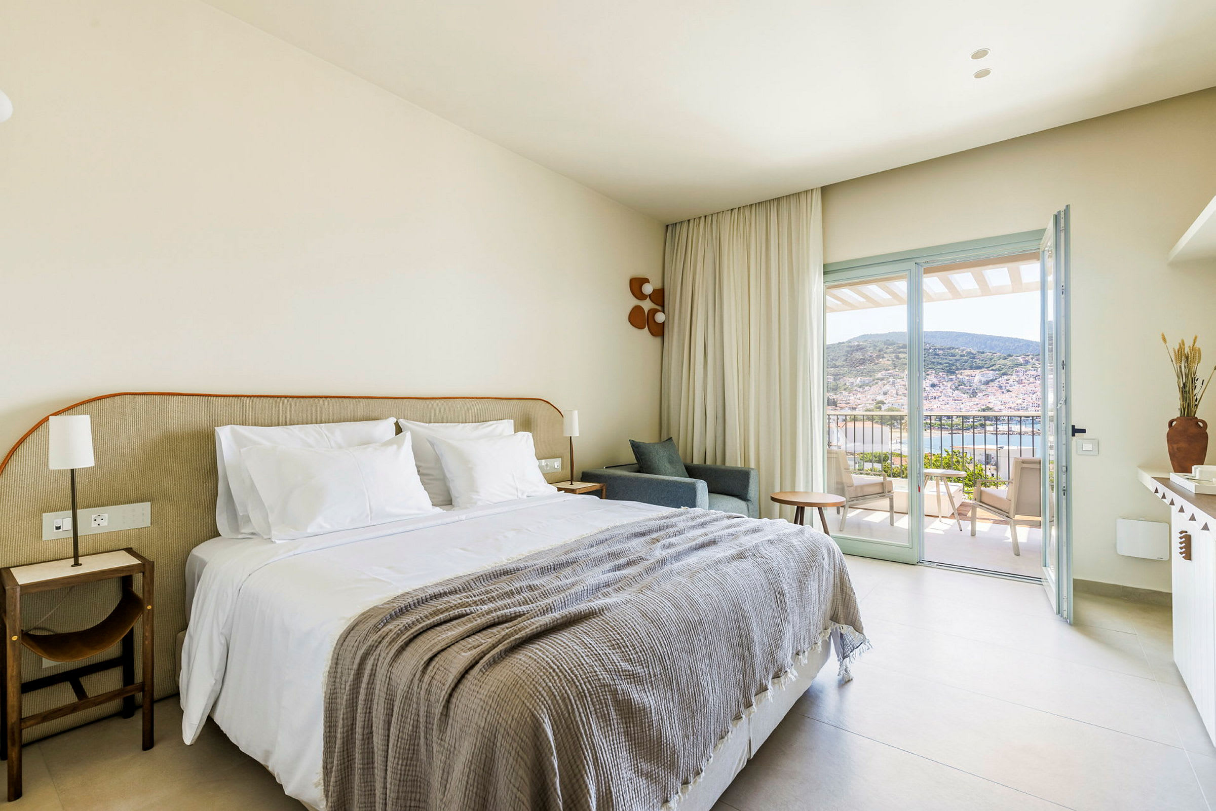Interiör av ett hotellrum med en dubbelsäng, balkong med utsikt över hav och kullar. (AI-genererad text)