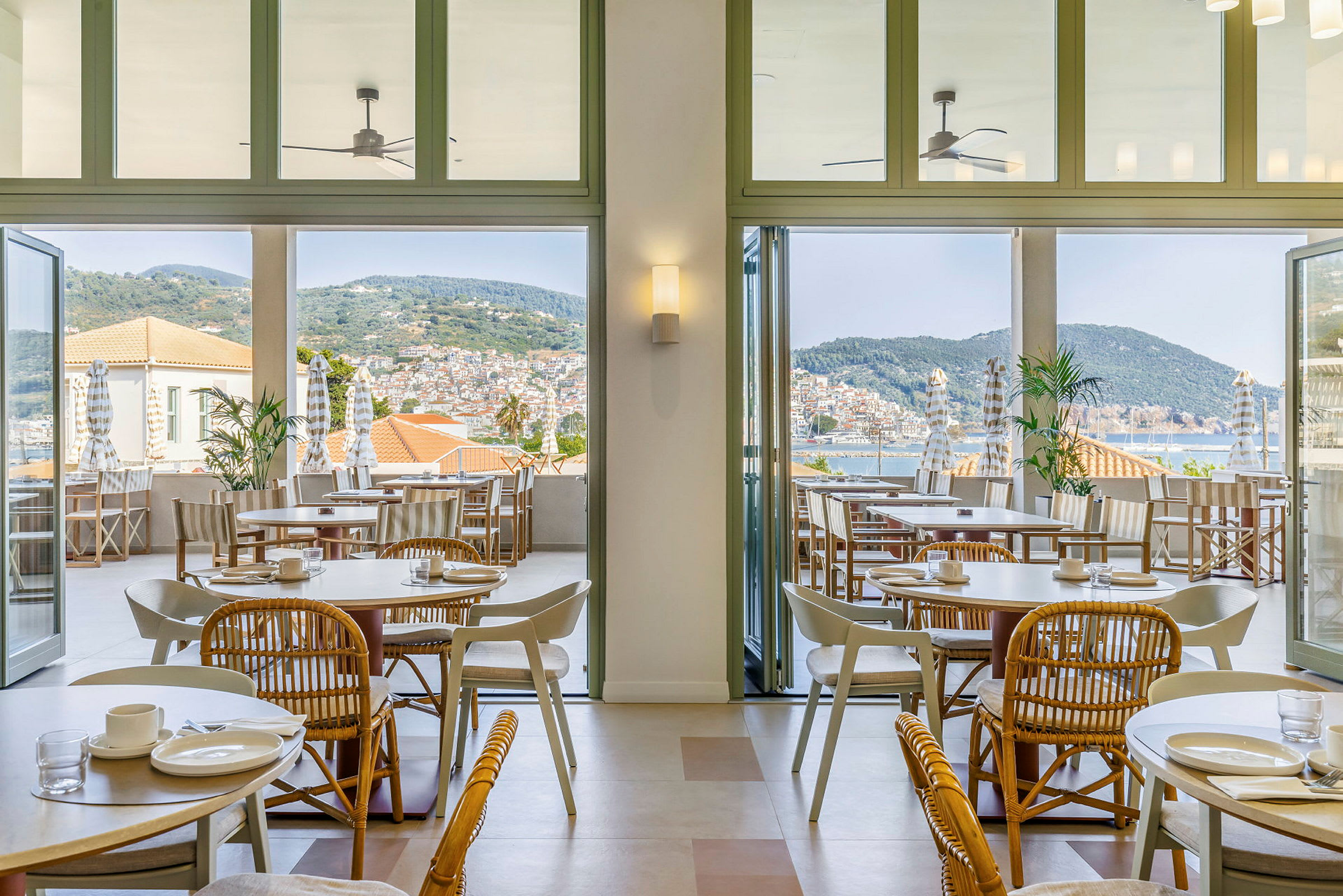 Bild av ett ljust och luftigt restaurangområde med bord och stolar inomhus samt en terrass med utsikt över havet och staden. (AI-genererad text)
