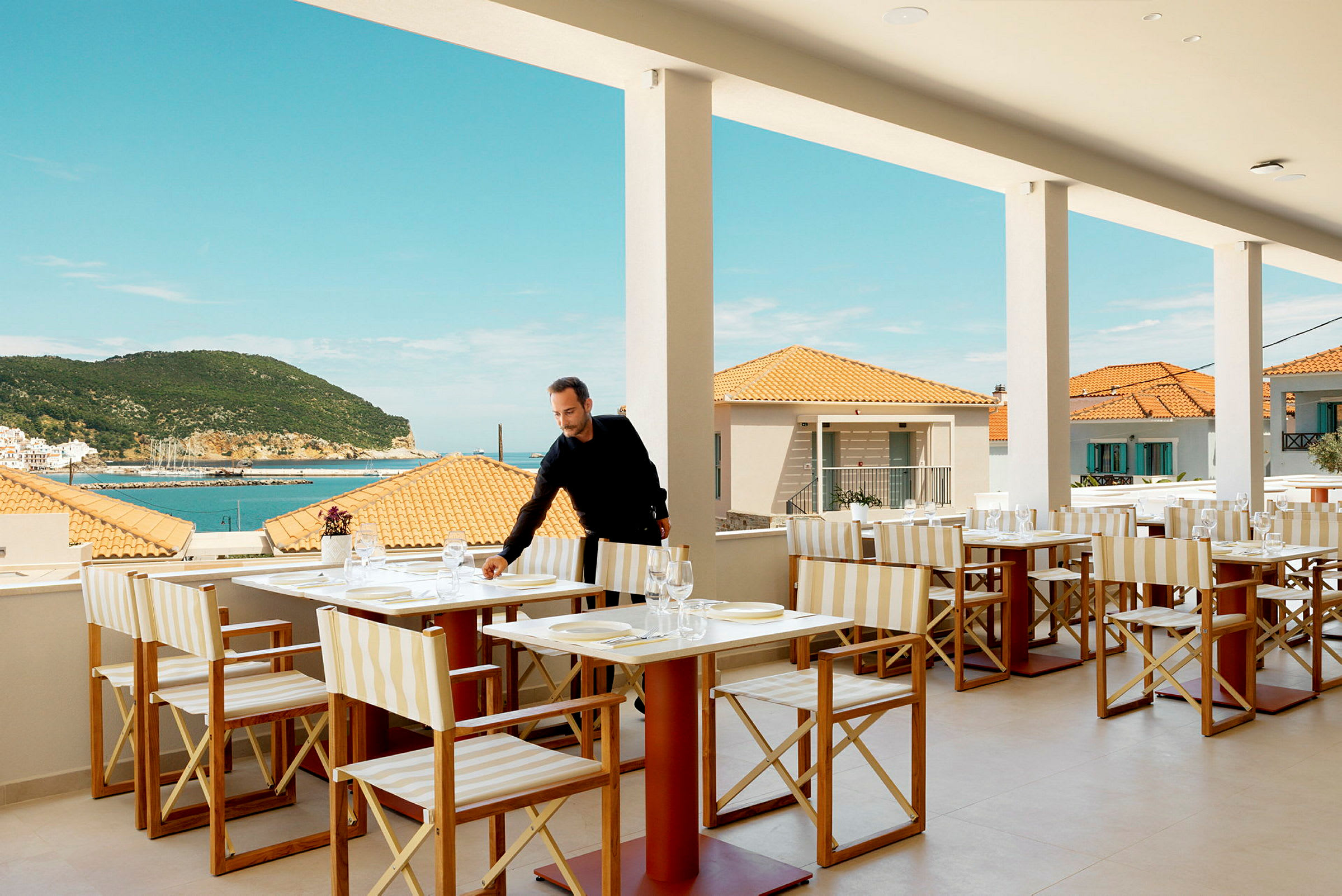 Restaurang med bordsuppsättning, utsikt över havet och terrass, mot bakgrund av terrakottatak och en grönskande bergskulle. (AI-genererad text)