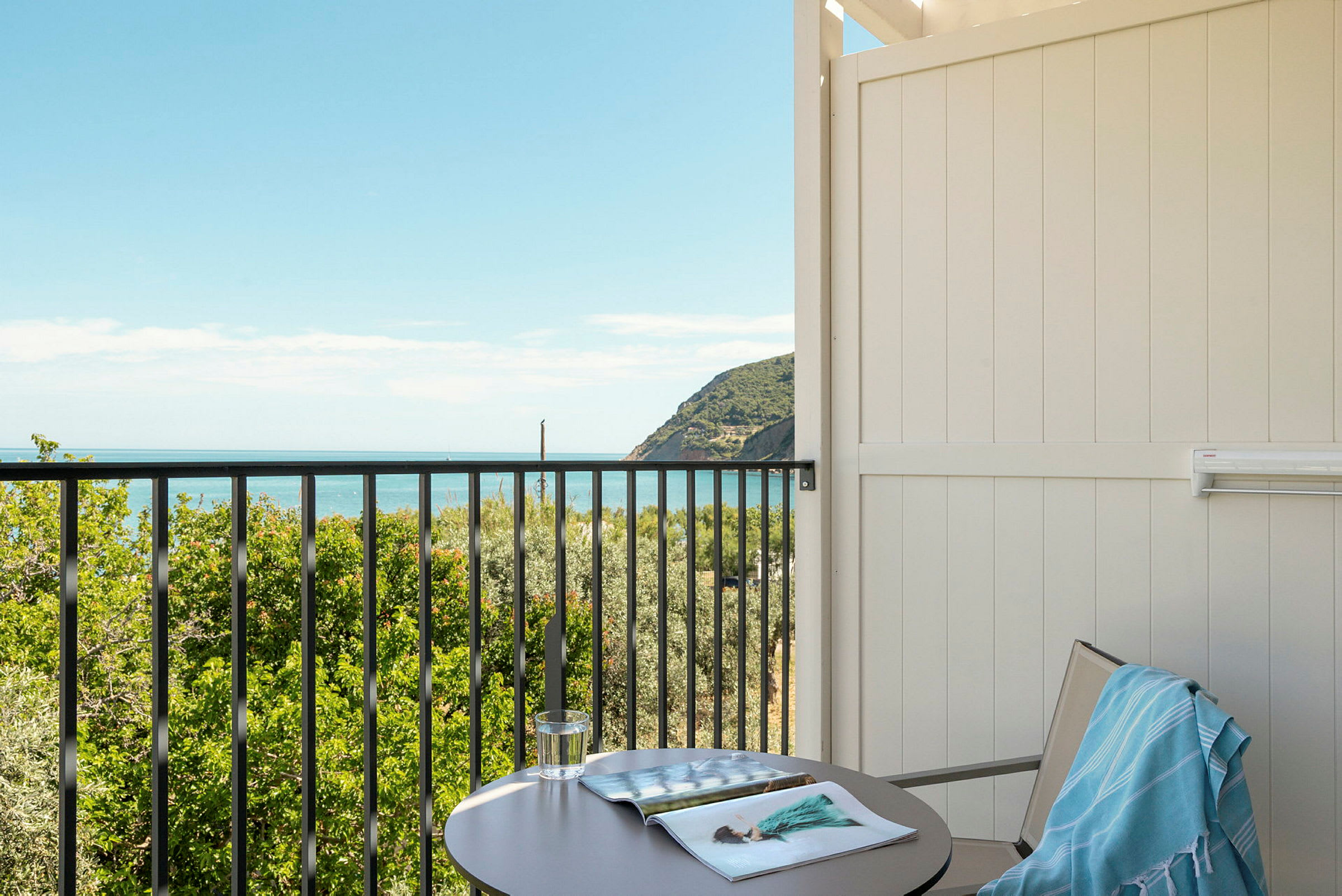 En balkong med utsikt över grönska och havet. På bordet ligger ett glas och en öppen tidning. (AI-genererad text)
