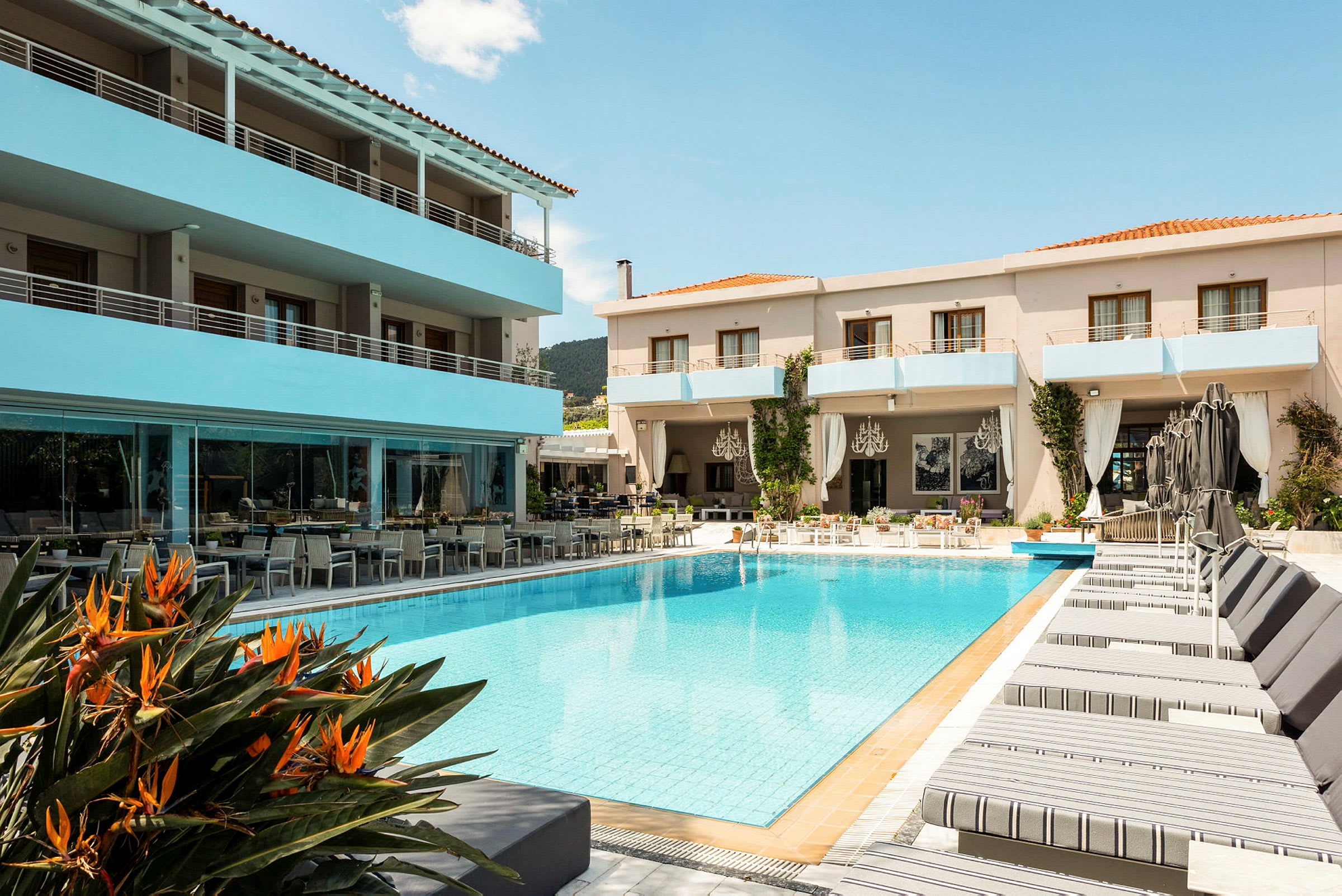 Ett hotell med en pool i förgrunden, omgiven av solstolar och några orangea blommor närmast bilden. Byggnaden har balkonger och en uteservering. (AI-genererad text)