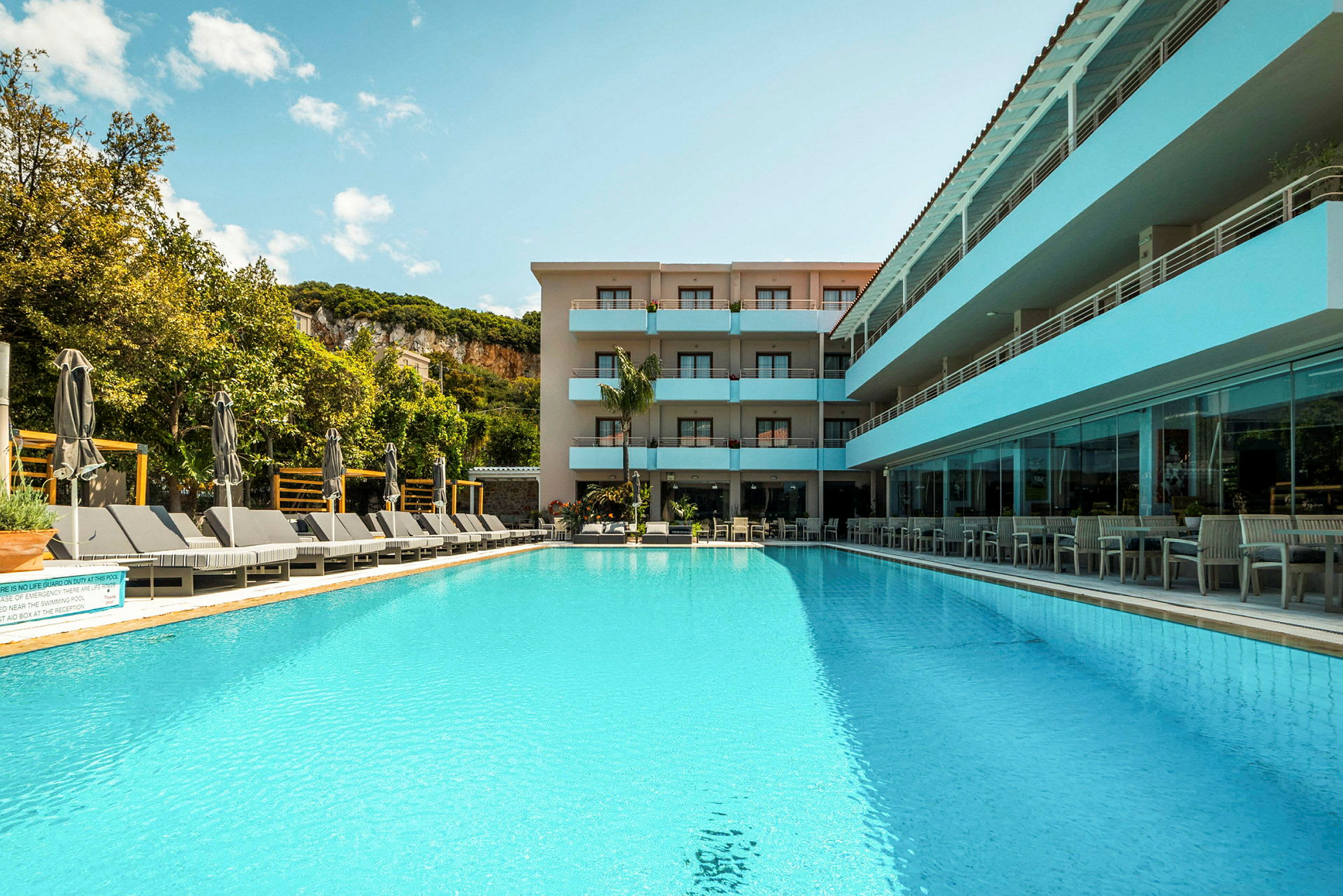 En utomhuspool framför ett hotell omgivet av grönska och klippor. (AI-genererad text)