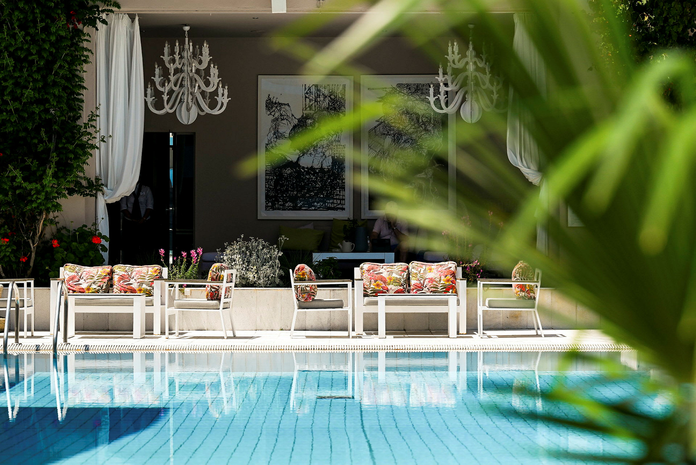 En utomhusmiljö med pool och sittgrupp med färgglada kuddar i förgrunden, omgiven av grönska. (AI-genererad text)