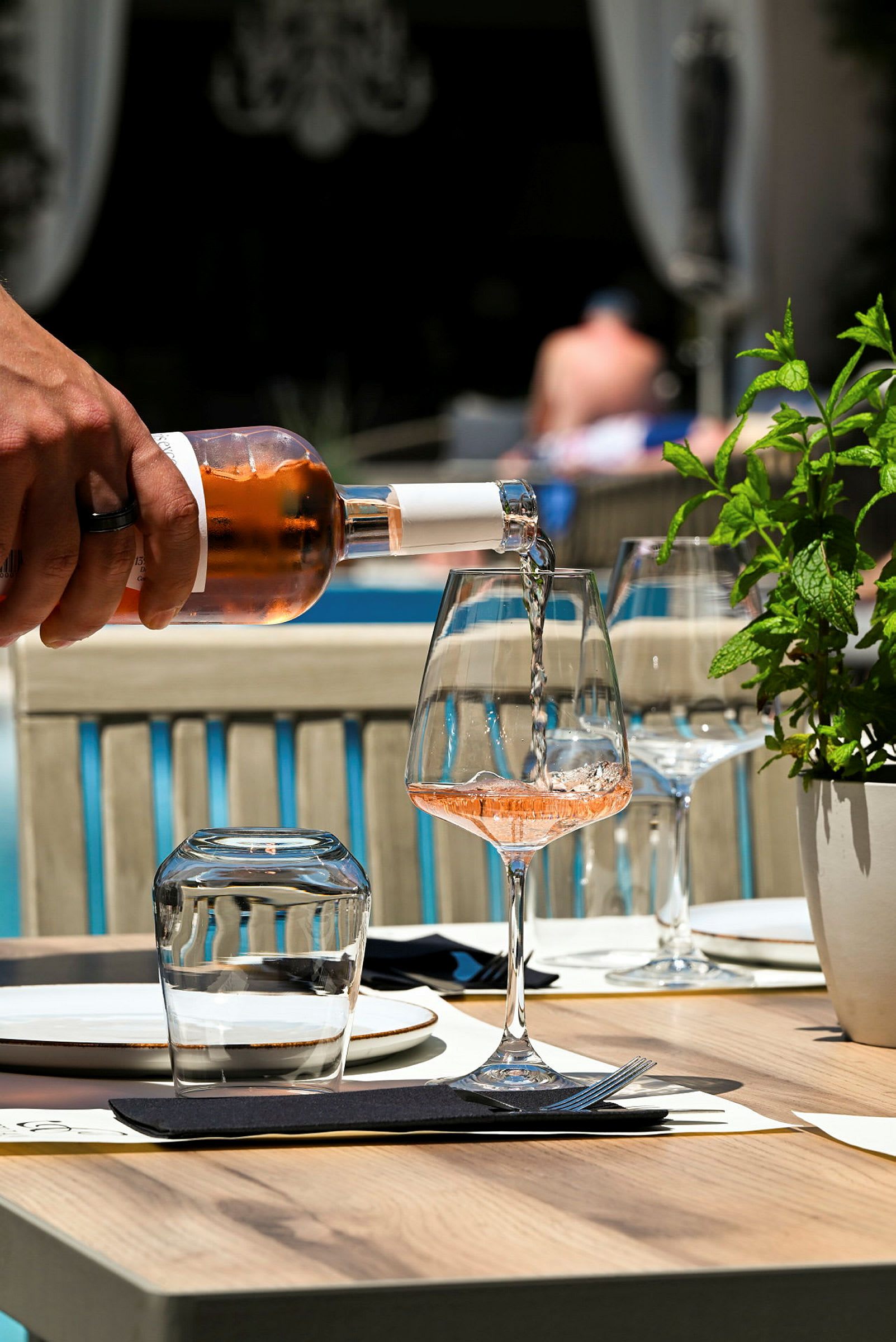 Ett vinglas fylls med rosévin vid ett dukat bord bredvid en grön växt. (AI-genererad text)