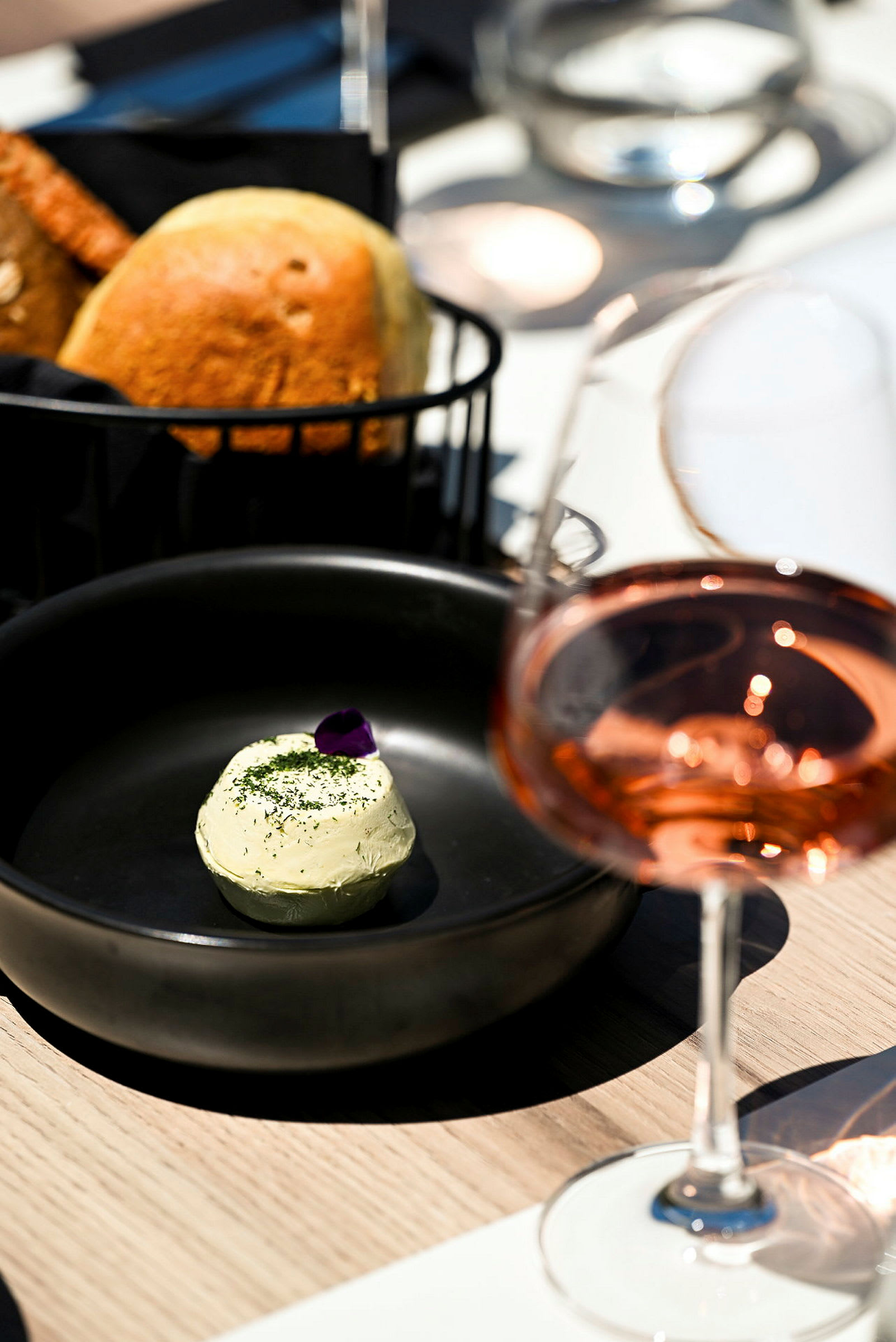 Ett svart fat med smör dekorerat med en blomma, ett bröd i en korg och ett glas rosévin på ett bord. (AI-genererad text)