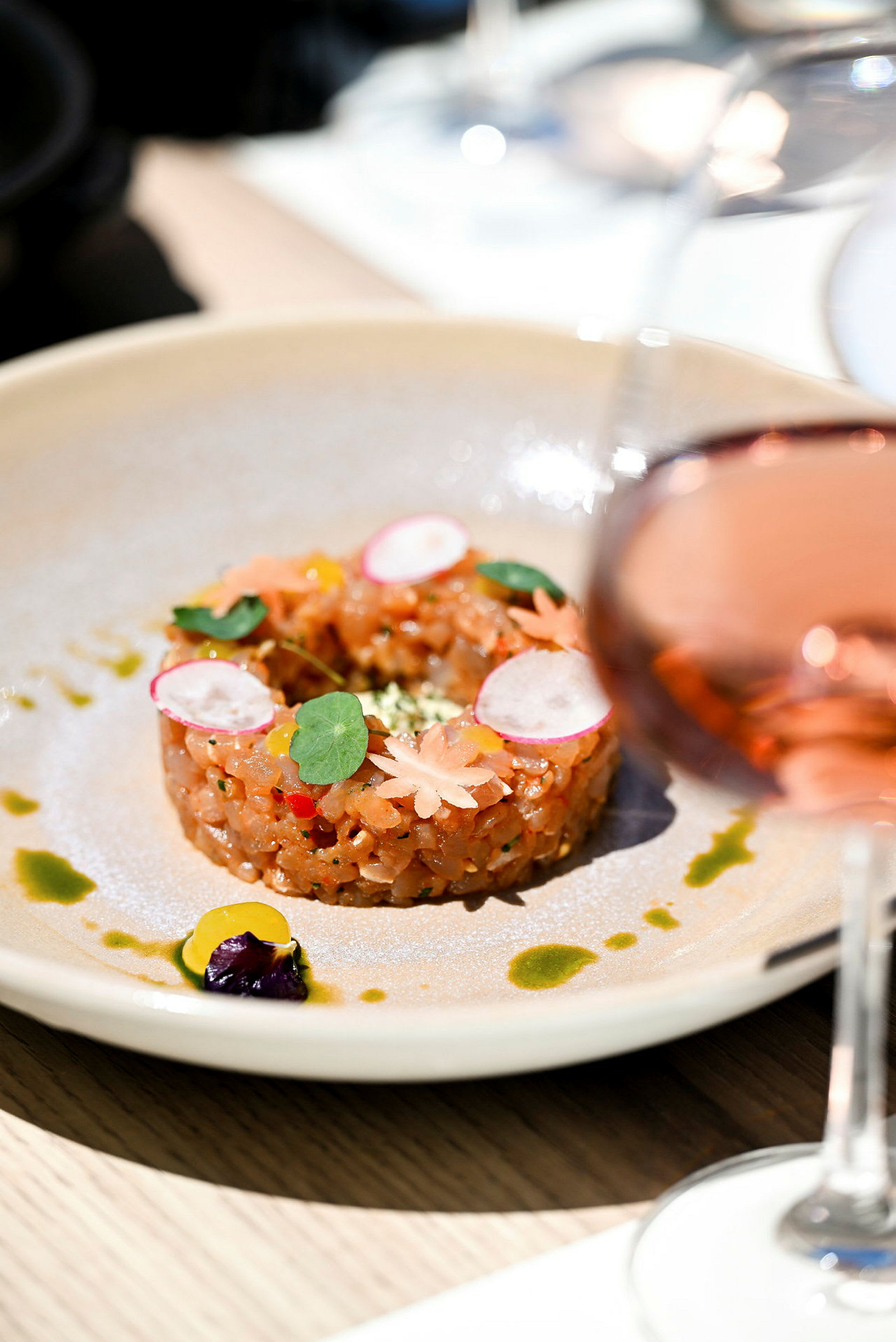 En tallrik med en dekorerad maträtt som ser ut som en tartar, omgiven av små såsdetaljer. Ett glas med en roséliknande dryck i förgrunden. (AI-genererad text)
