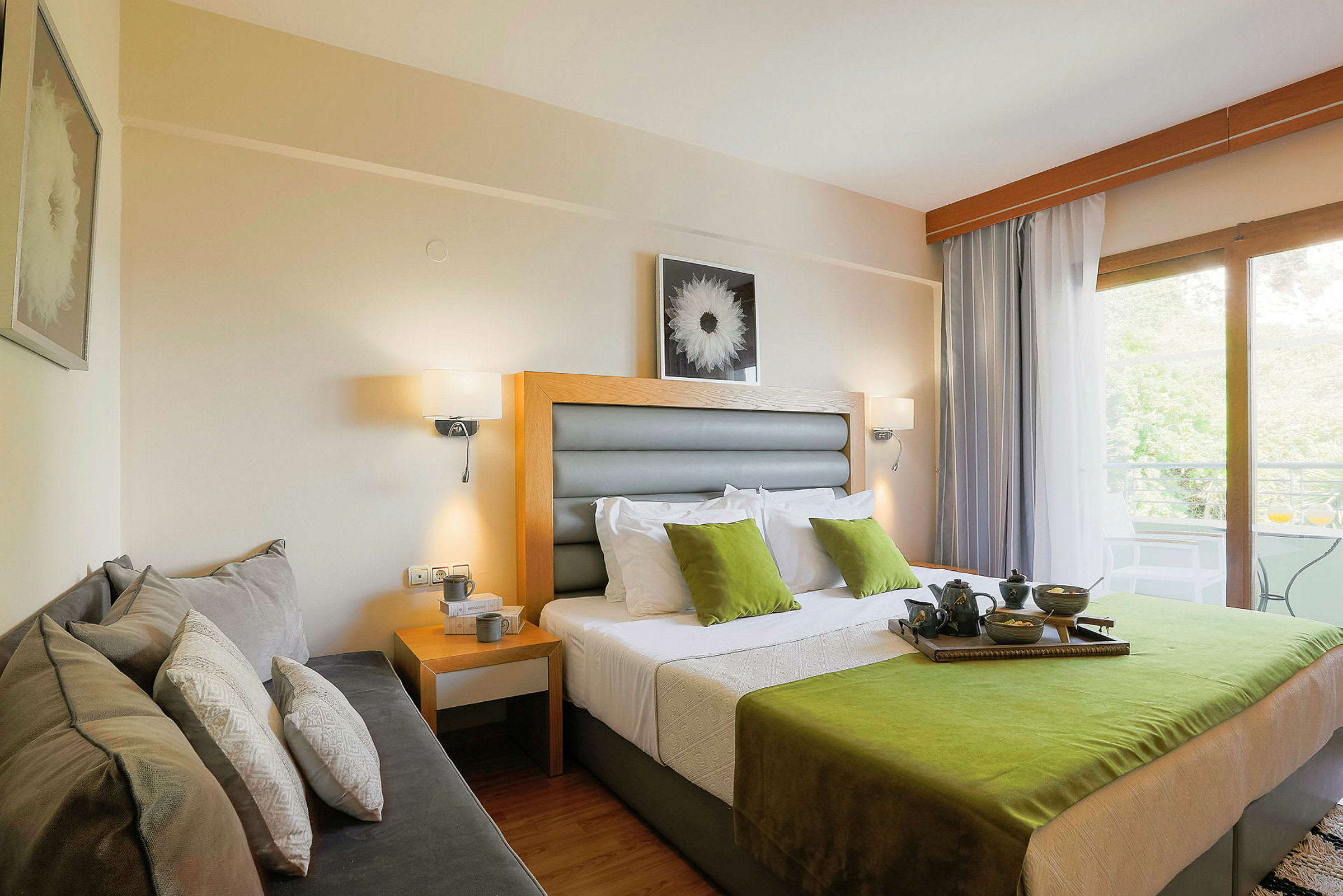 Ett hotellrum med en dubbelsäng med gröna och vita detaljer, en soffa på sidan, och ett fönster som leder till en balkong med utsikt. (AI-genererad text)