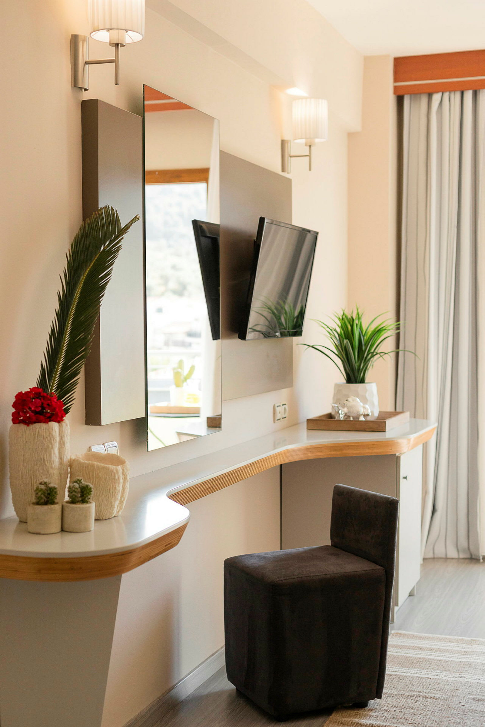 Ett modernt hotellrum med skrivbord och spegel, en liten stol och dekorativa växter. (AI-genererad text)