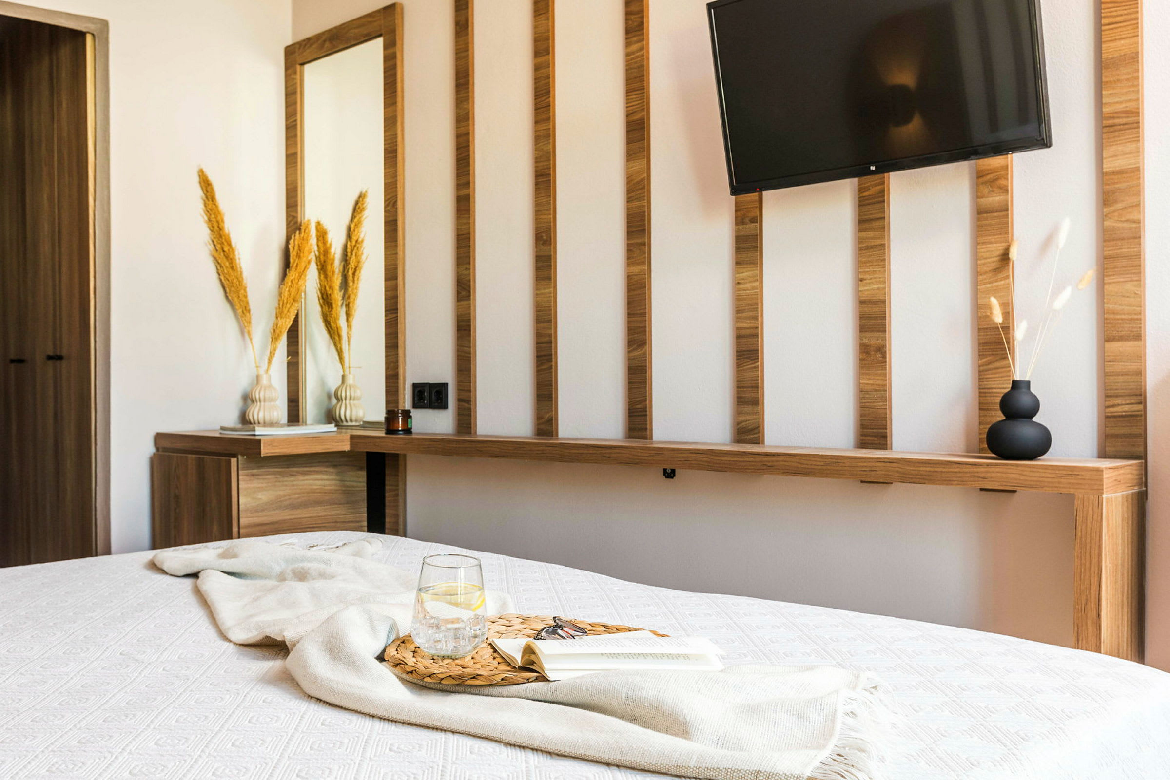 En modernt designad hotellrum med ett vitt sängöverkast, ett glas med vatten och en bok på en träbricka, samt en vägg med träpaneler, en spegel och en TV. (AI-genererad text)
