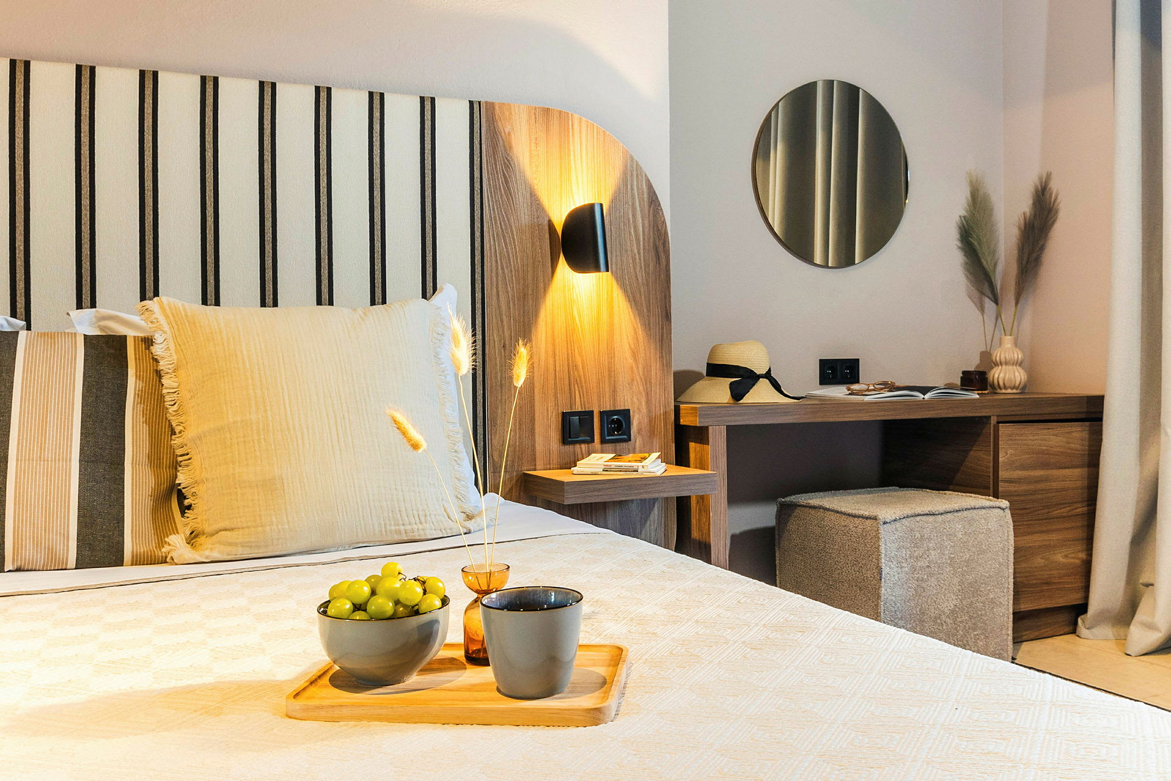 Ett modernt hotellrum med trädekor, en bädd med dekorativa kuddar, ett skrivbord med spegel och dekorativa föremål, och gröna druvor på ett fat. (AI-genererad text)