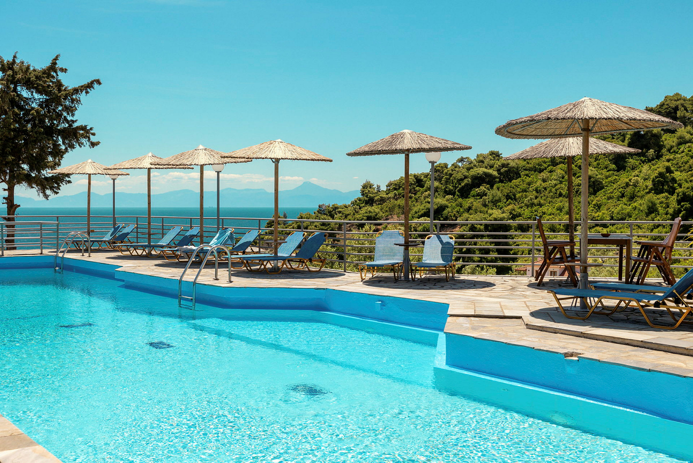 Pool med blå vatten, omgiven av solstolar och parasoll med utsikt över grönska och hav. (AI-genererad text)