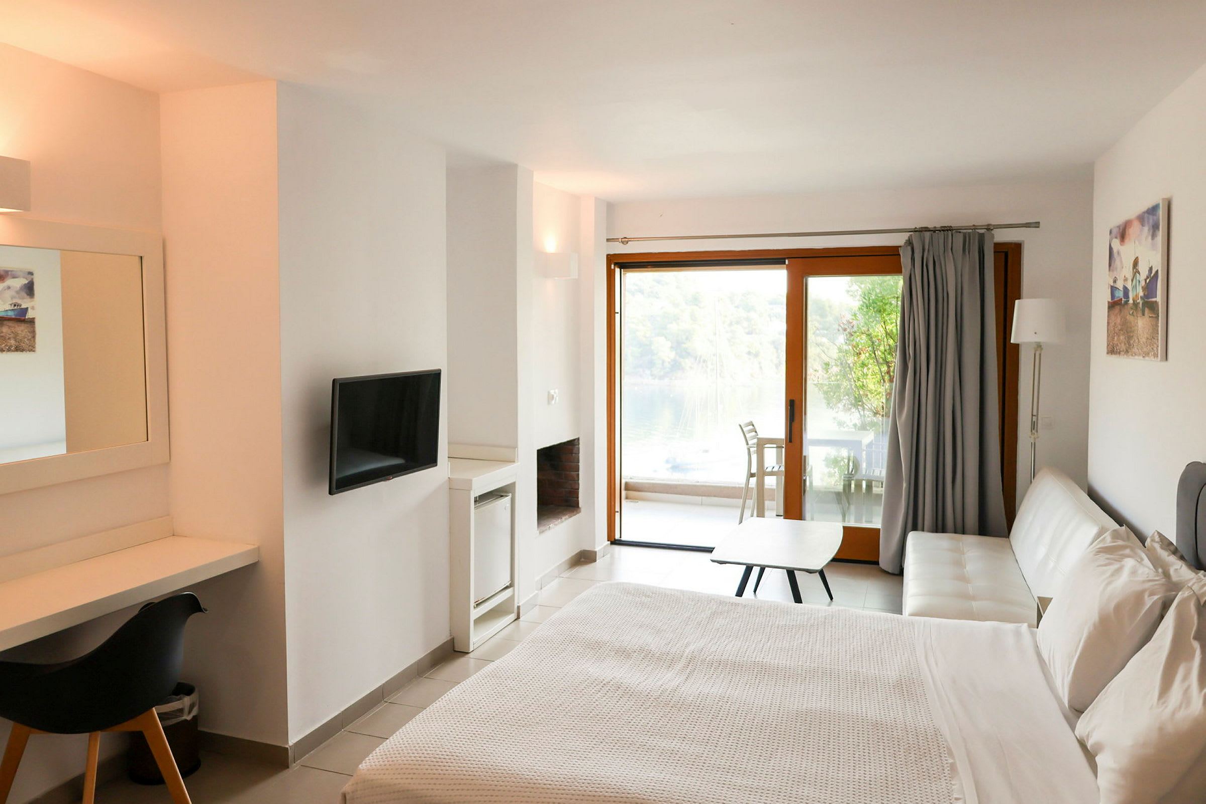 Ett modernt hotellrum med en dubbelsäng, skrivbord, TV och en vit soffa. I bakgrunden syns en glasdörr som leder till en möblerad altan med utsikt över natur och vatten. (AI-genererad text)