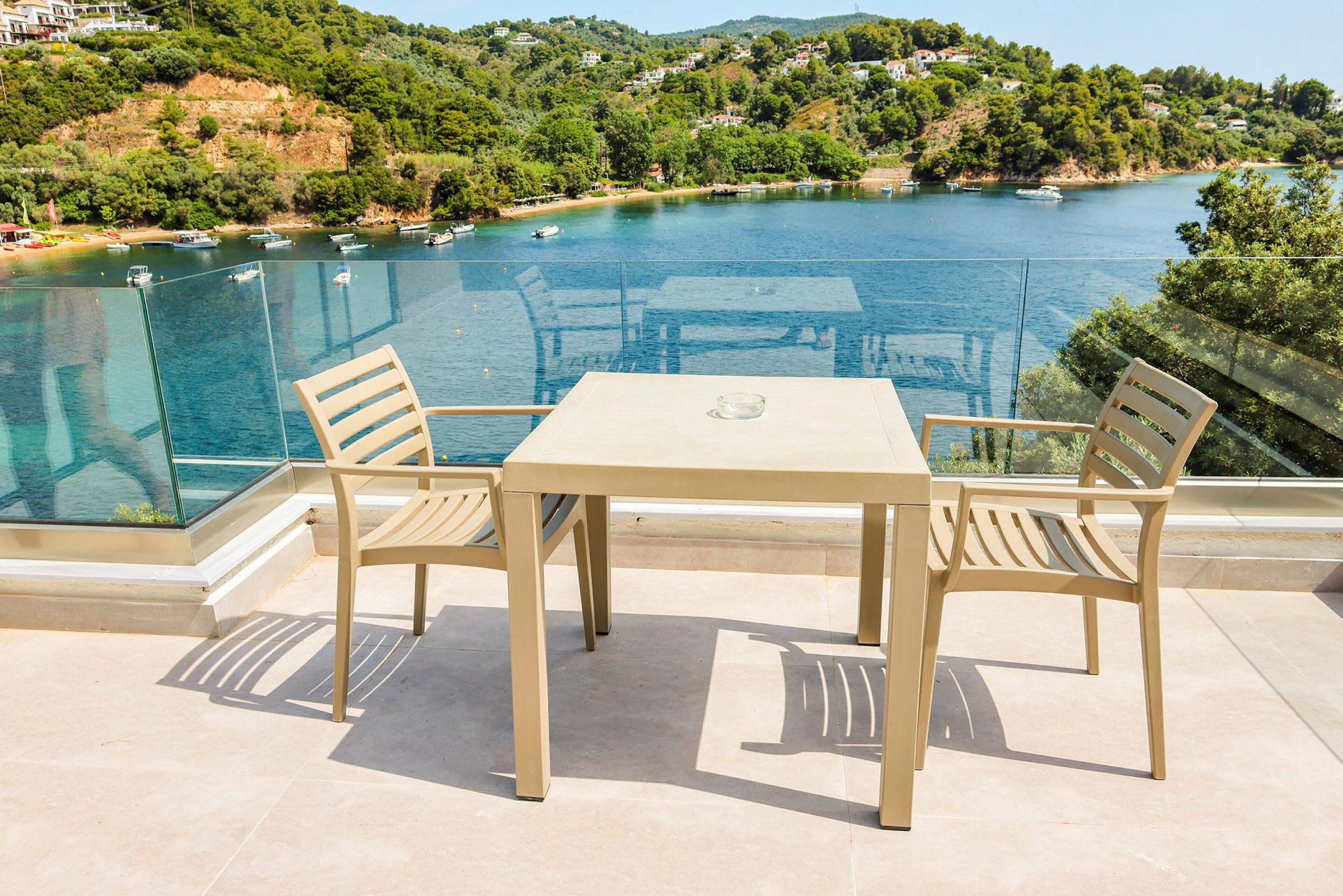 Två beige stolar och ett bord på en terrass med glasräcke och utsikt över en vik med båtar och grönska. (AI-genererad text)
