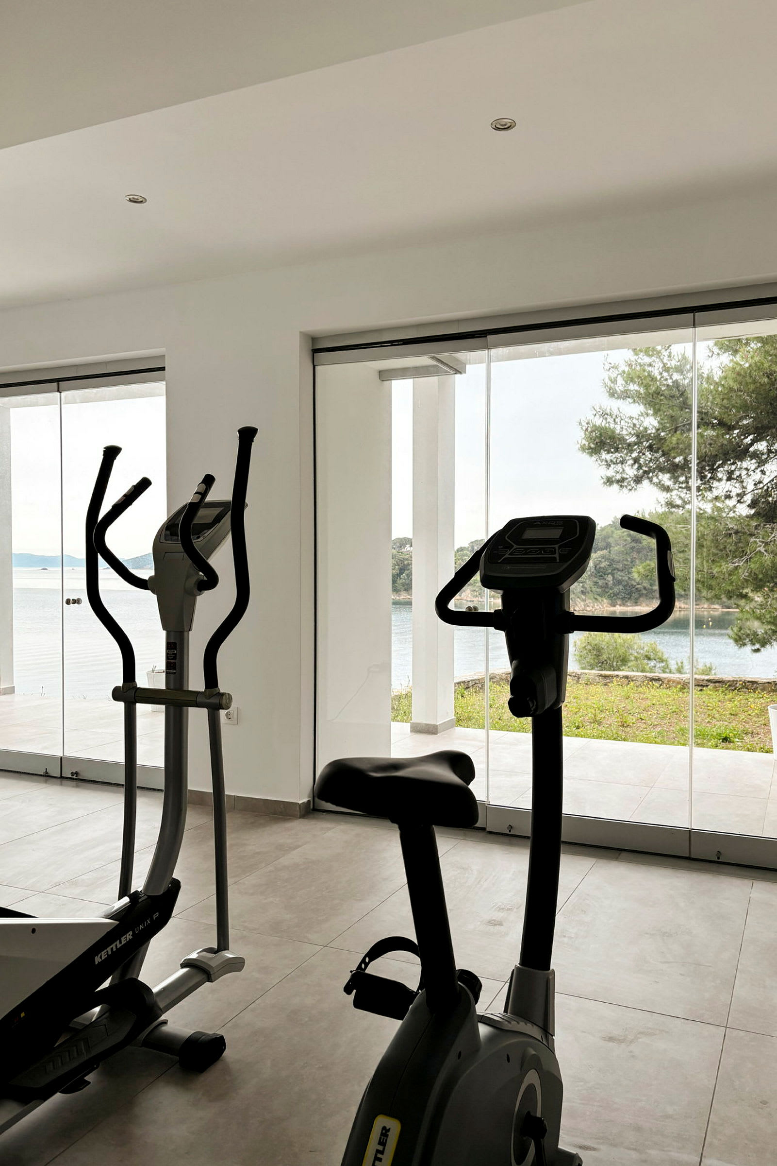 Ett gym med träningsmaskiner, inklusive motionscykel och crosstrainer, med stora fönster som visar en utsikt över natur och hav. (AI-genererad text)