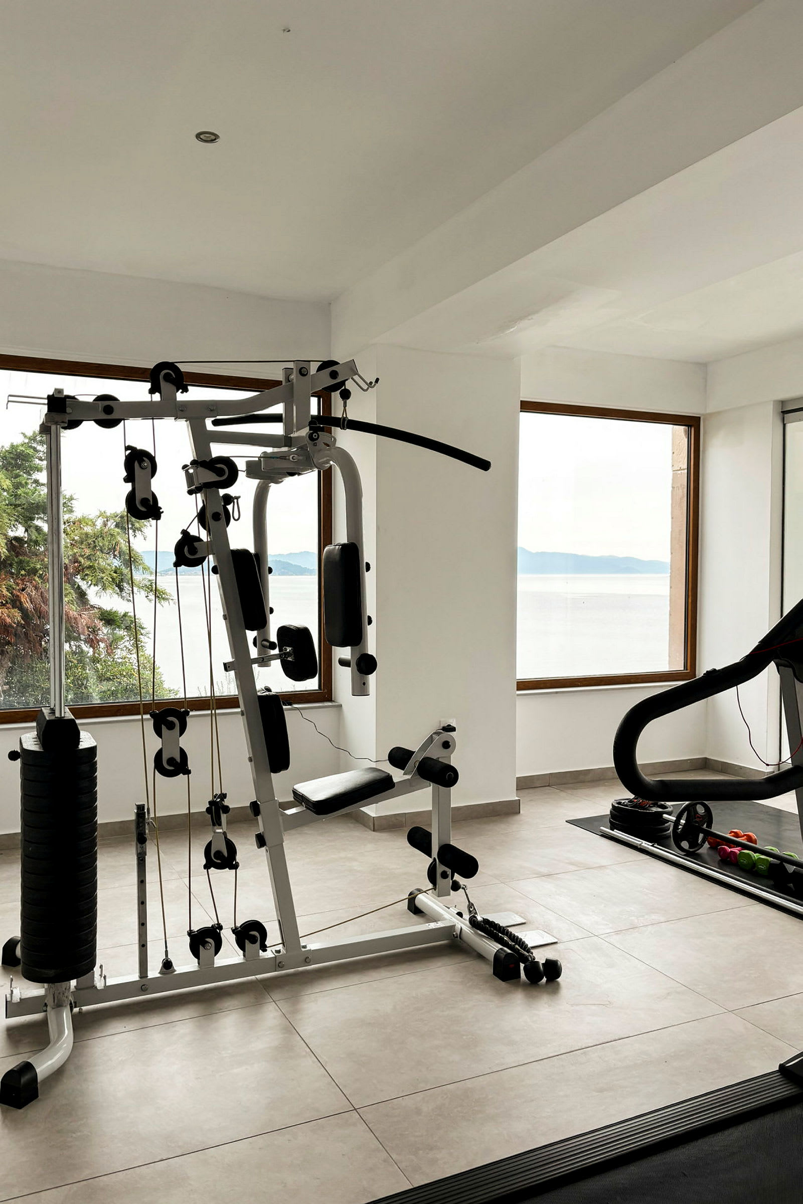 Ett gym med träningsutrustning inklusive ett multigym och en löpband, med fönster som vetter mot en havsutsikt. (AI-genererad text)