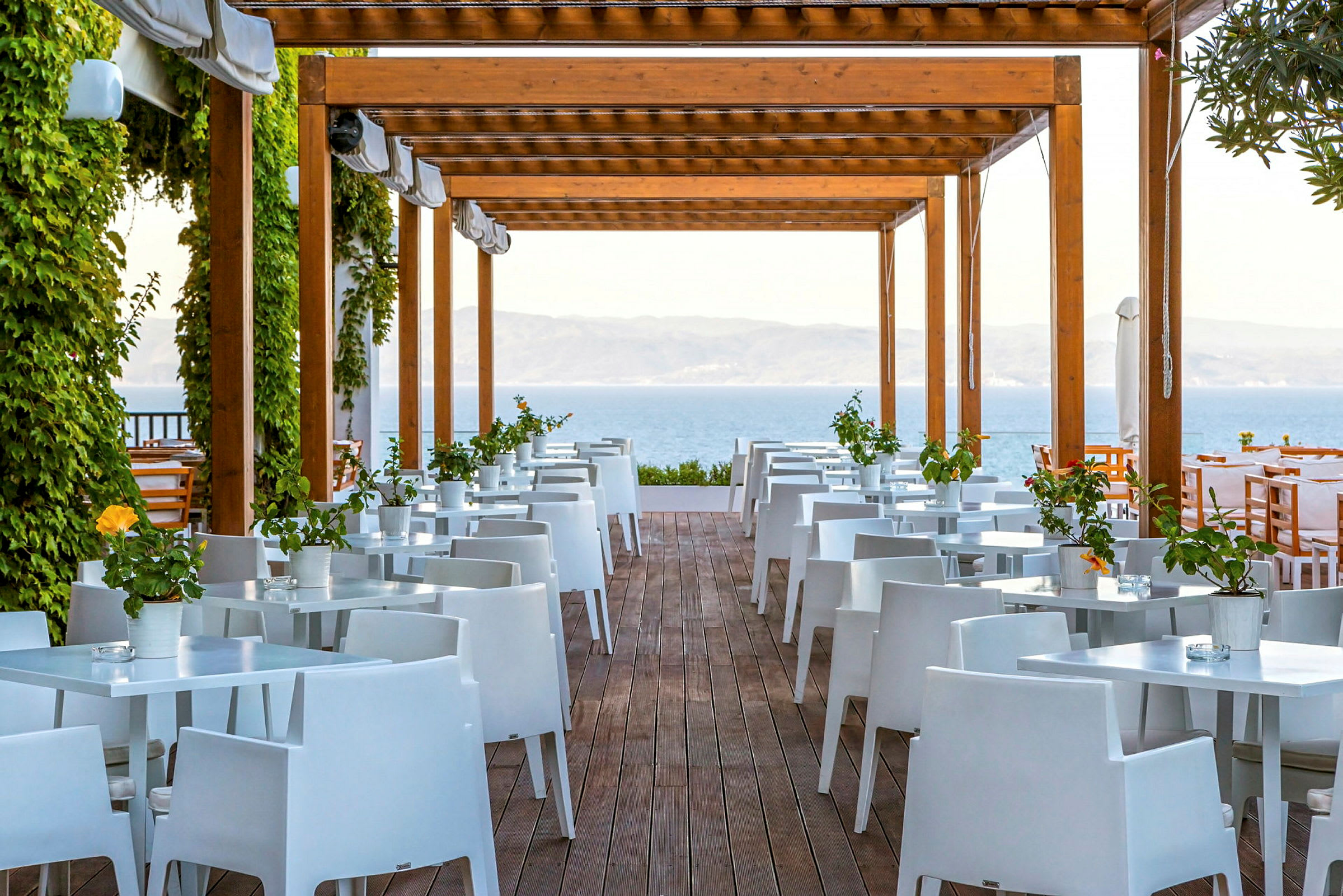 Utomhusrestaurang med vita bord och stolar under träpergola, omgivna av grönska. (AI-genererad text)