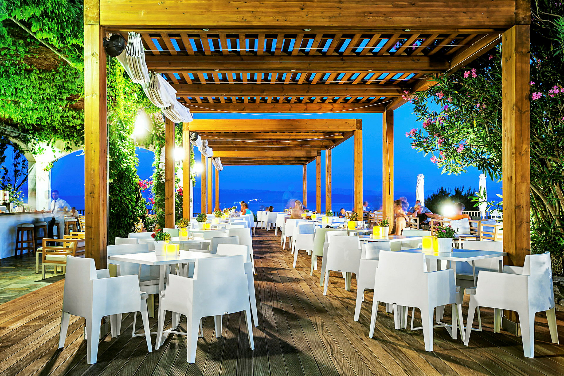Utomhusrestaurang med träpergola, vita stolar och bord, omgiven av grönska och kvällsbelysning. (AI-genererad text)