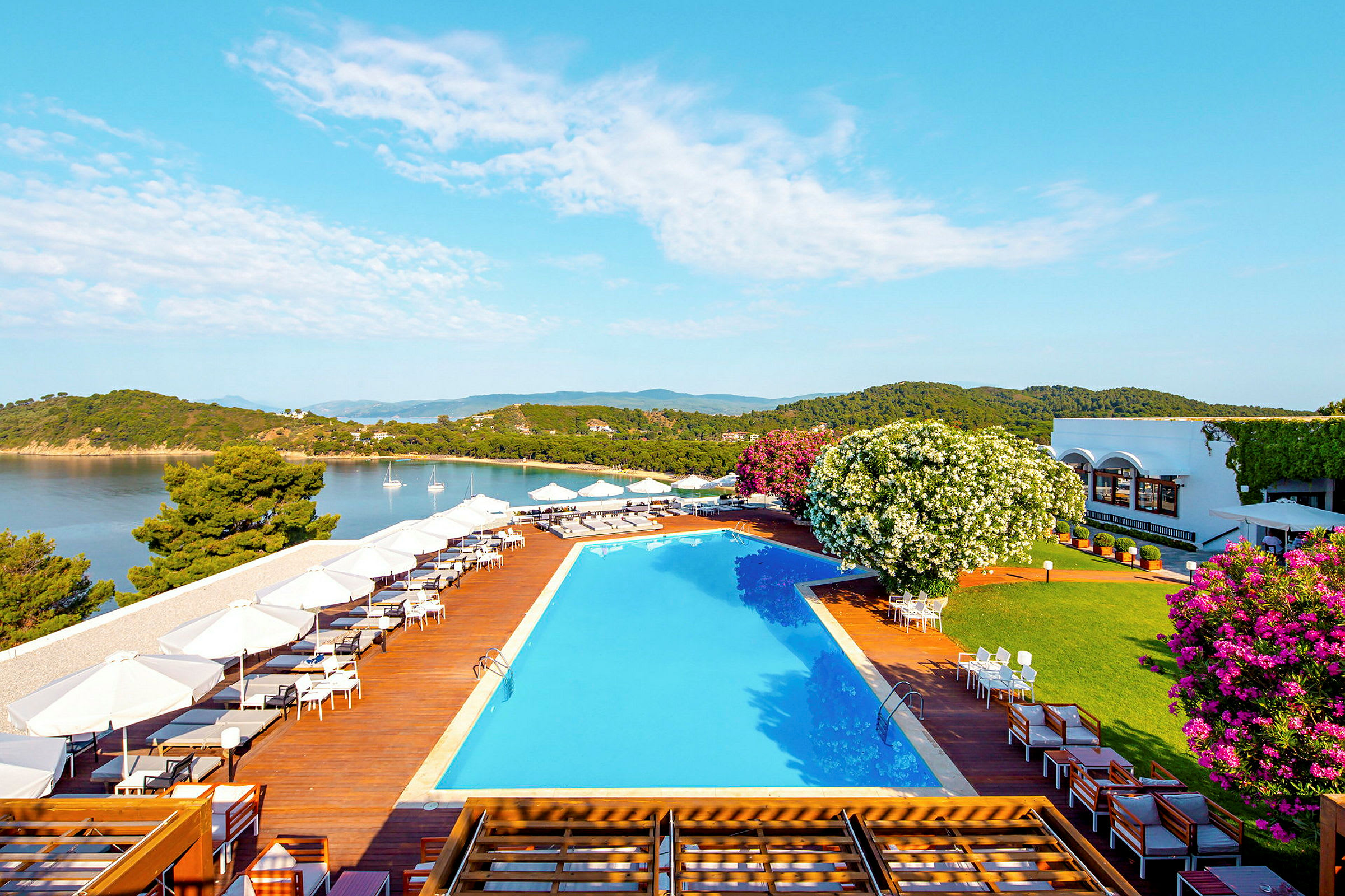 Exteriör av en pool- och uteplatsområde bredvid havet, med grönskande vegetation och byggnader i bakgrunden. (AI-genererad text)