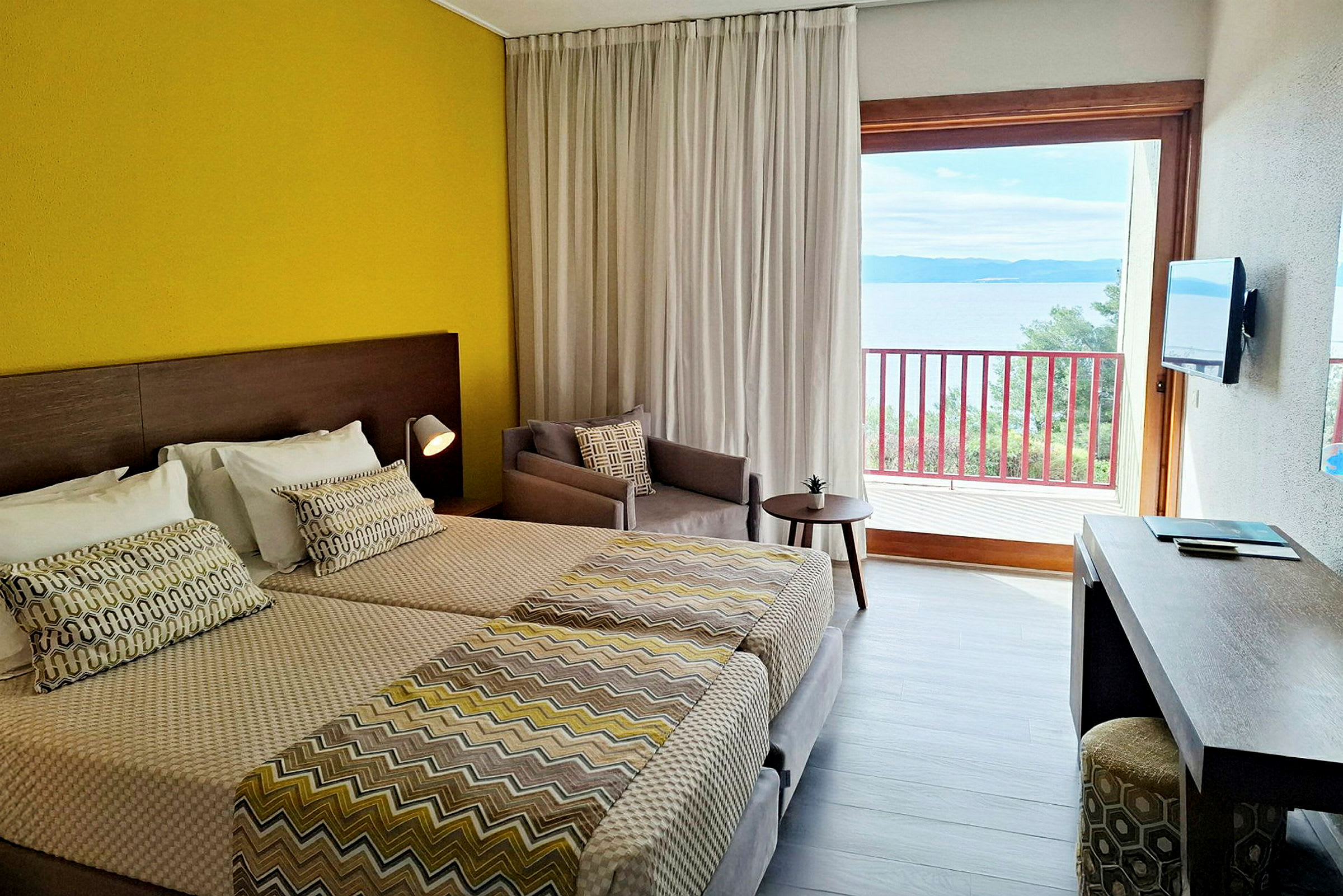 Ett hotellrum med två sängar, en gul vägg, ett skrivbord, en beige fåtölj och en balkong med utsikt över havet. (AI-genererad text)