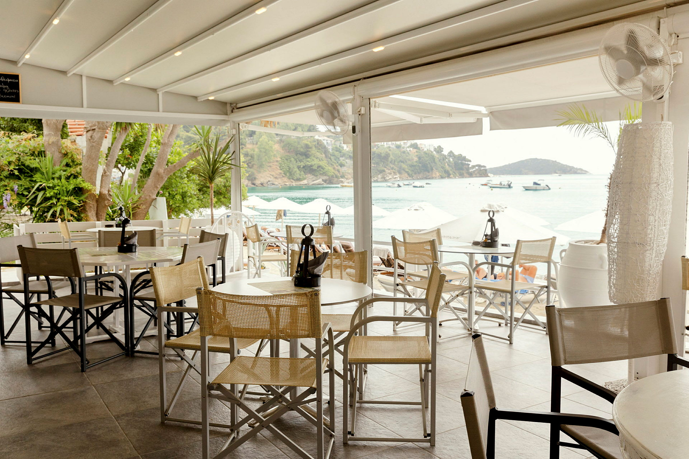 En restaurang med utsikt över havet, med bord och stolar samt parasoller i bakgrunden. (AI-genererad text)