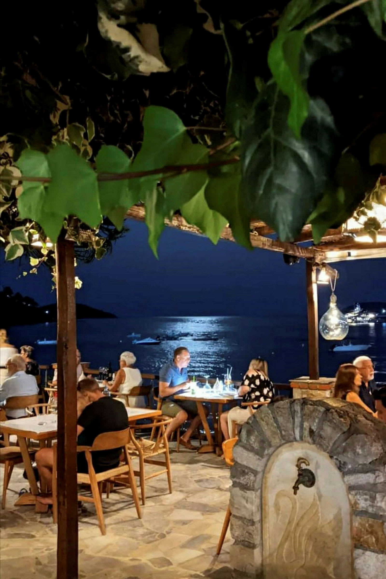 Uteservering vid havet med belysning och båtar synliga i bakgrunden. (AI-genererad text)