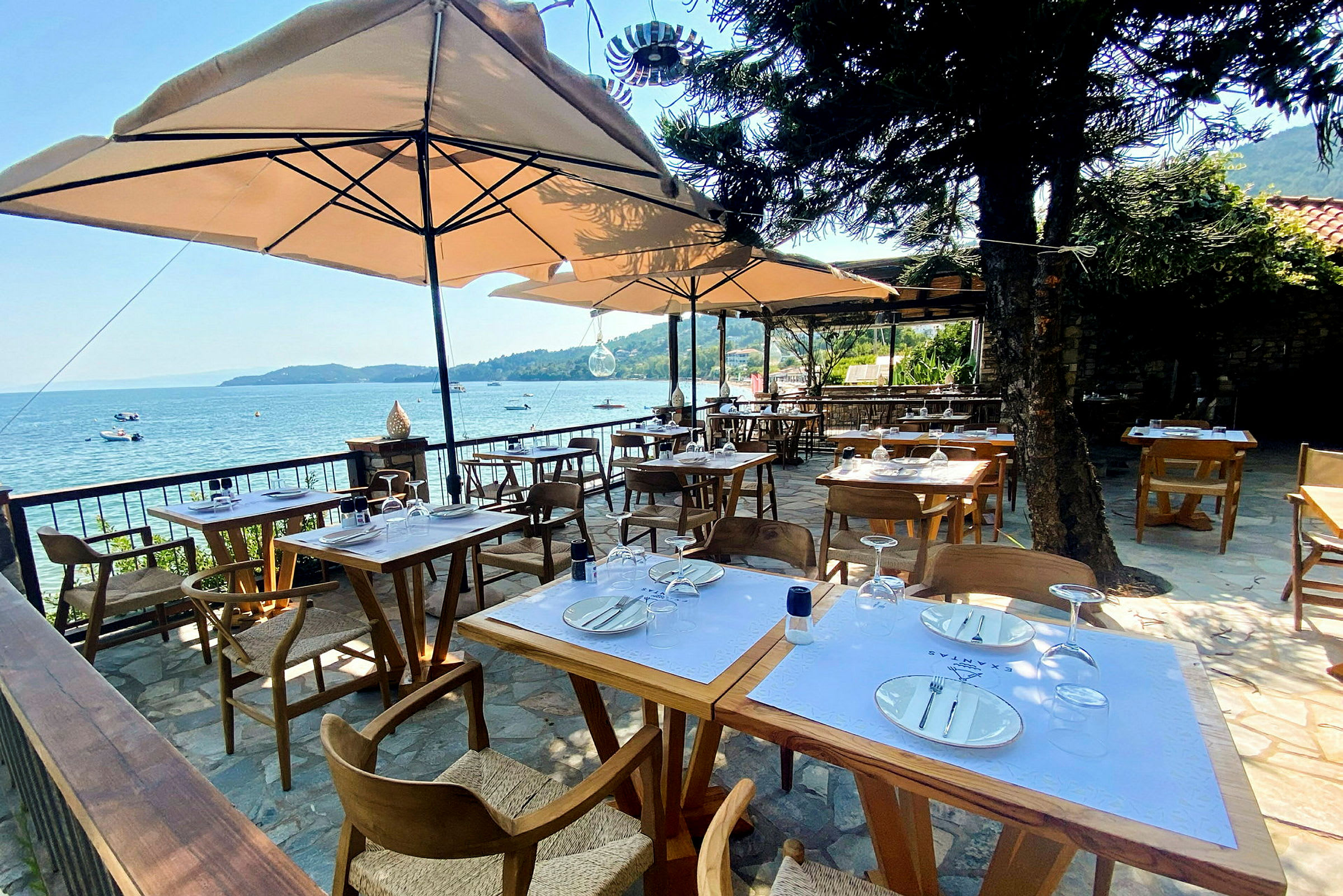 Bild på en utomhusrestaurang med bord och stolar under parasoller, med utsikt över havet i bakgrunden. (AI-genererad text)
