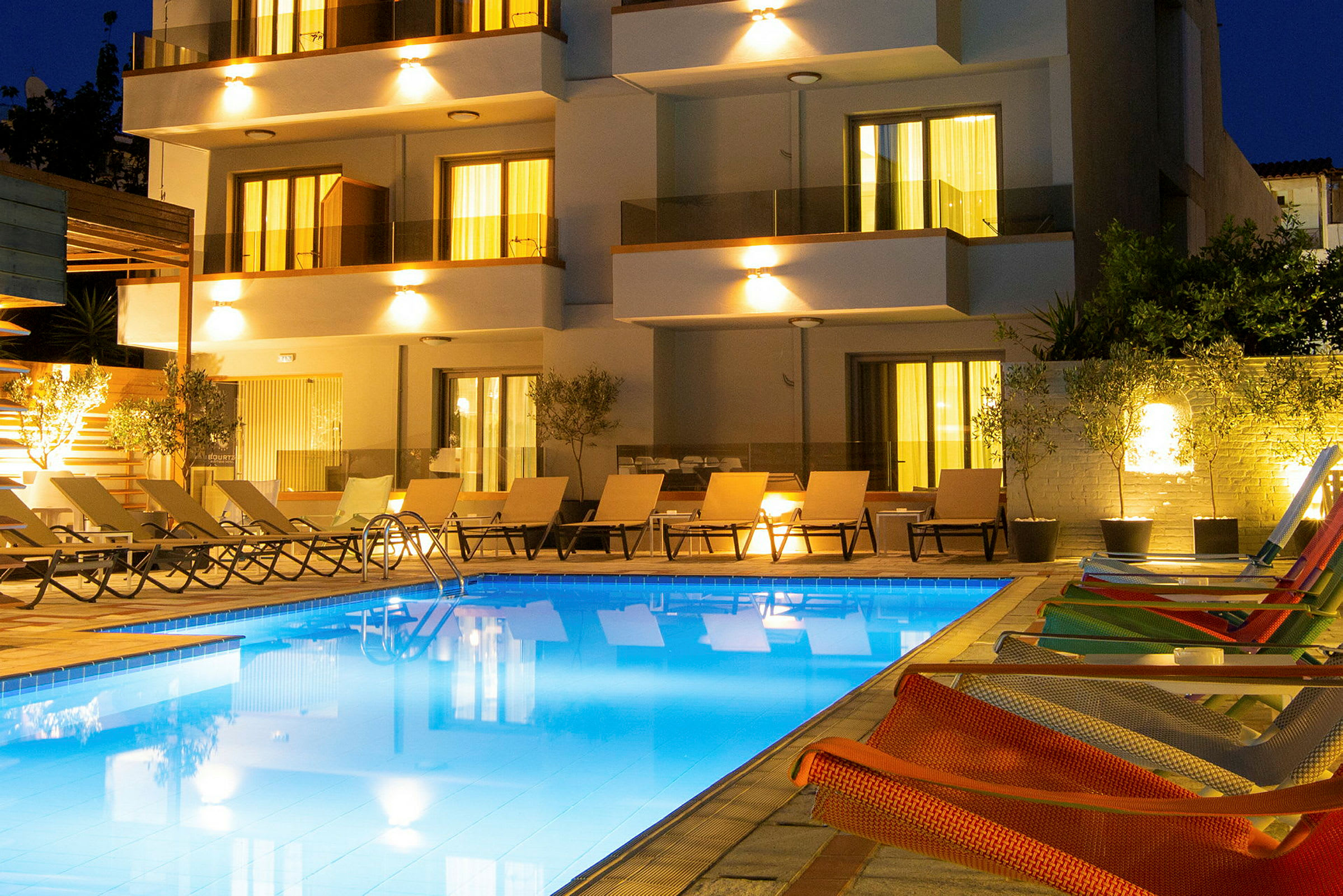 En upplyst pool med solstolar och en modern byggnad i bakgrunden under kvällen. (AI-genererad text)