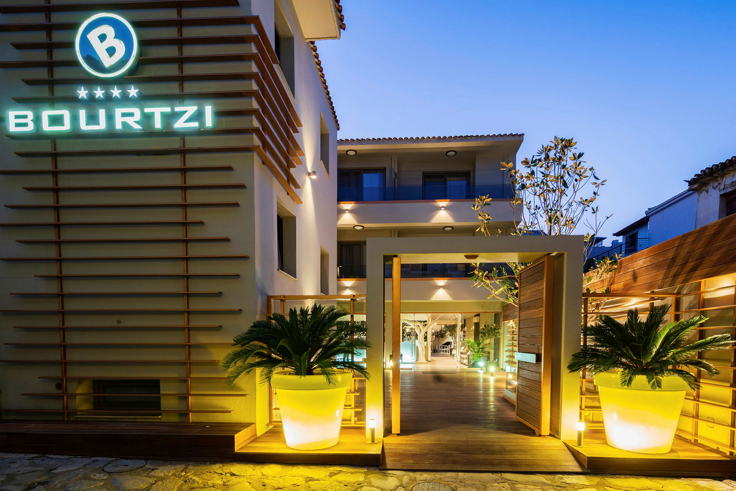 Entrén till ett hotell med namnet Bourtzi, upplyst i skymningen. Två stora plantor i ljusa krukor flankeras ingången. (AI-genererad text)