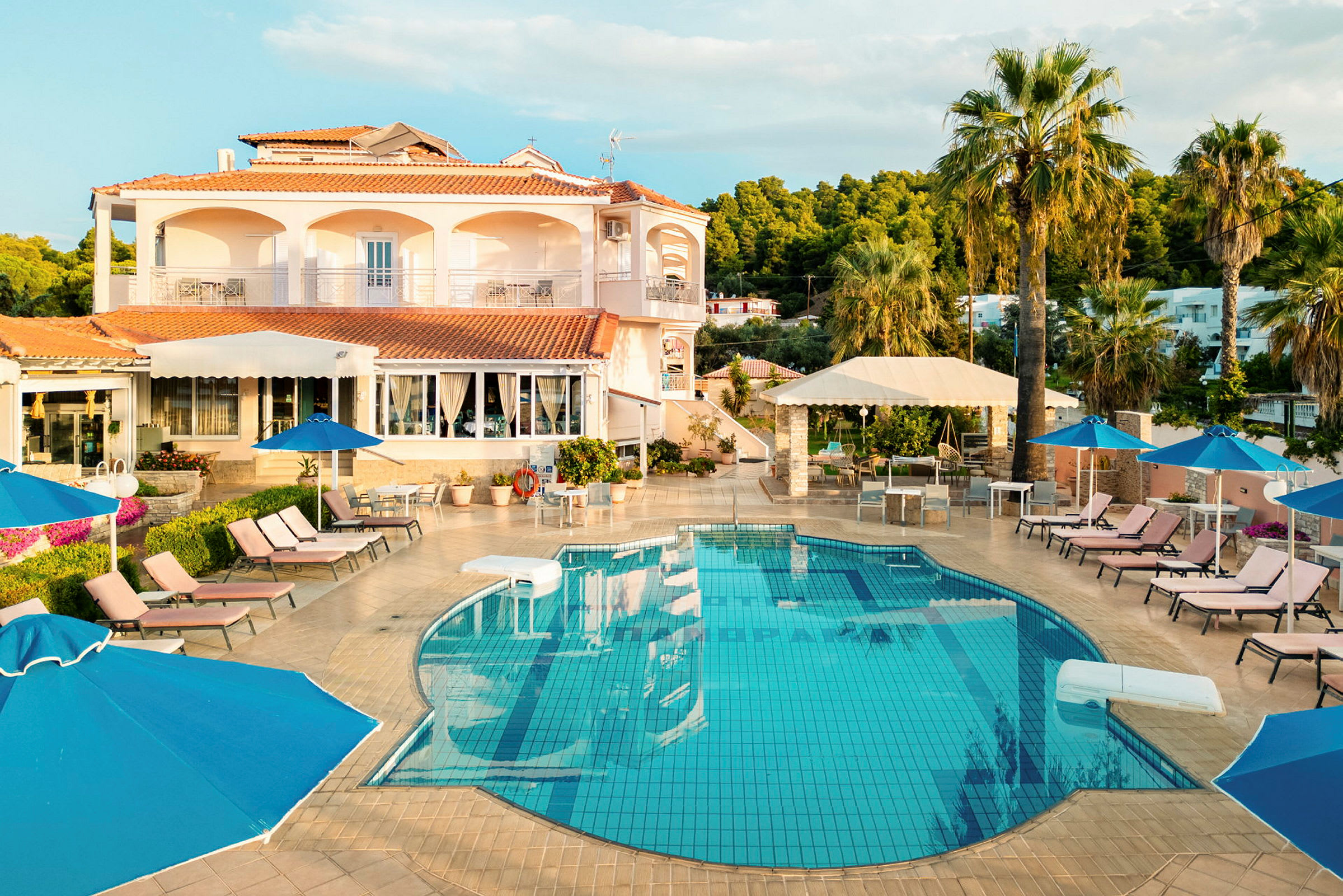 Bild på en utomhuspool vid ett hotell med solstolar, parasoller och palmer runtom samt en byggnad i bakgrunden. (AI-genererad text)
