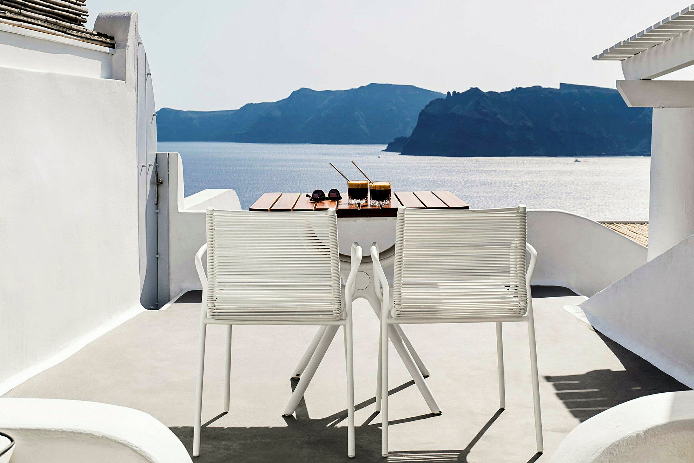 Ett bord med två drycker och ett par solglasögon står på en terrass med två vita stolar. Utsikten visar havet och landskap i bakgrunden. (AI-genererad text)