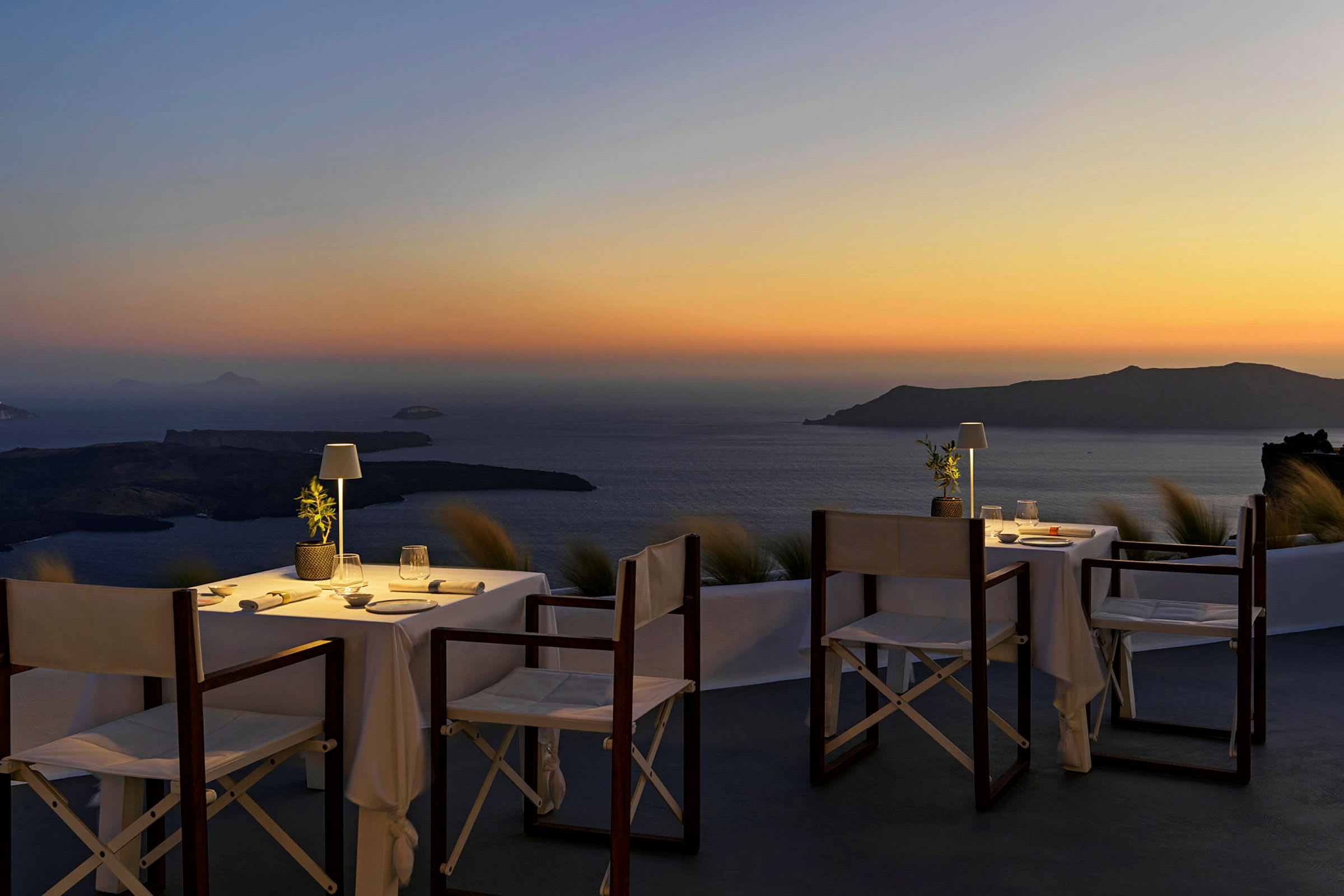 Två dukade bord med stolar på en terrass, med utsikt över havet och en ö vid solnedgång. (AI-genererad text)