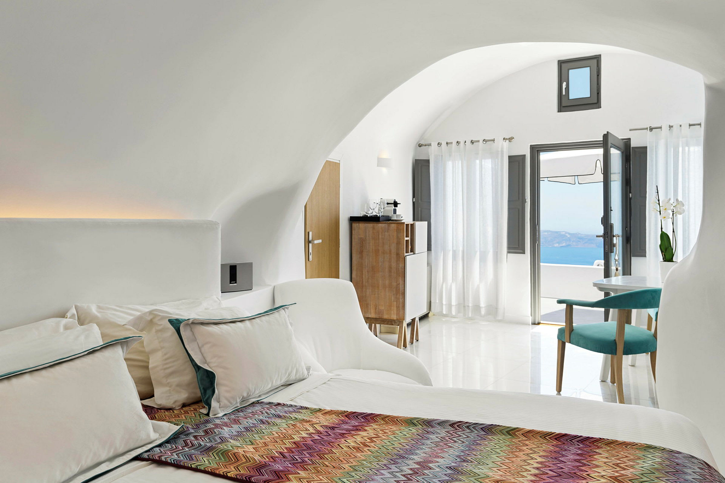 Ett ljust och modernt hotellrum med en säng, färgglad pläd, sittgrupp och stora fönster som vetter mot en utsikt över havet. (AI-genererad text)