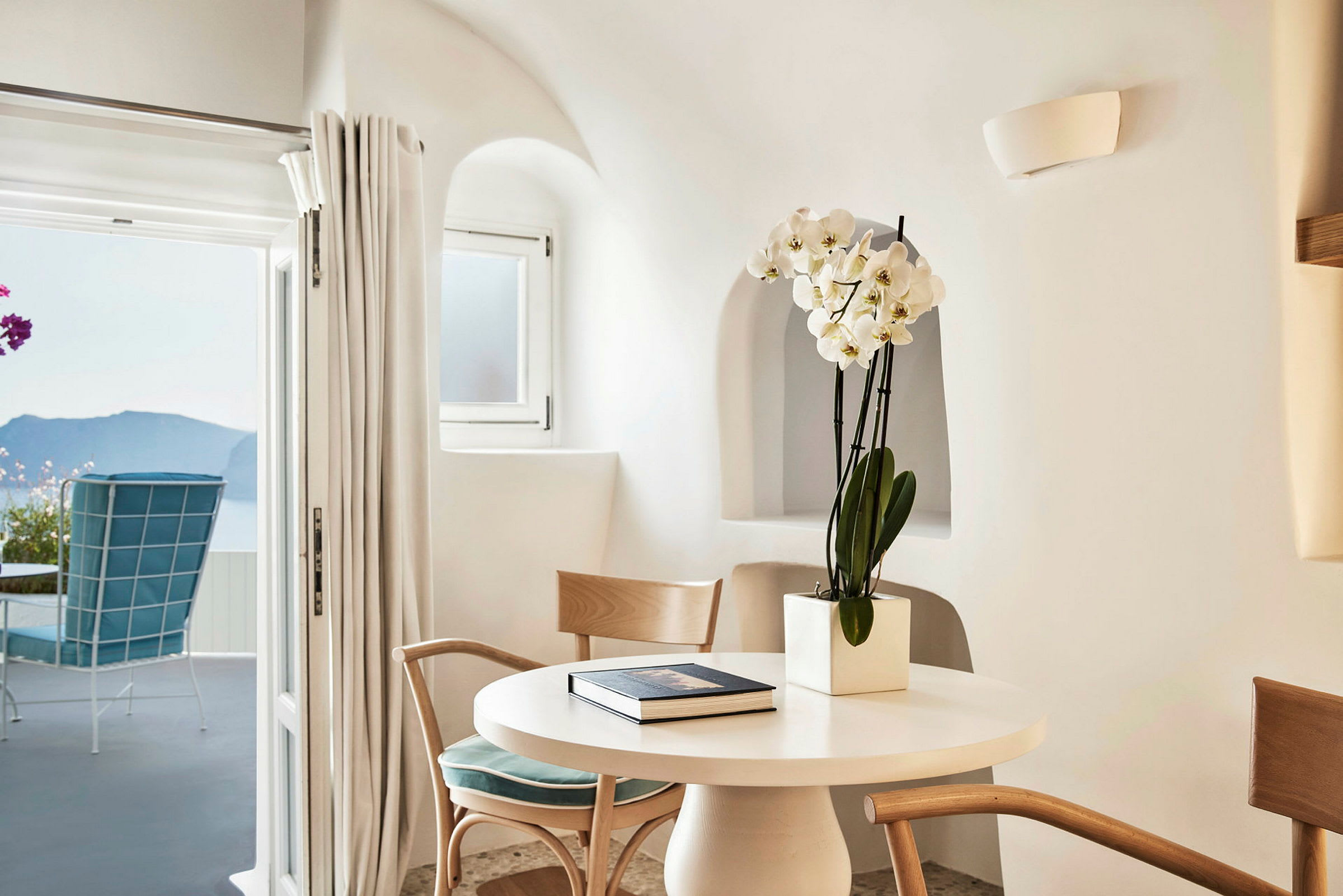 Rum i ljus och modern stil med runda fönster, blommor i kruka på bordet och en utsikt mot havet genom en öppen dörr. (AI-genererad text)