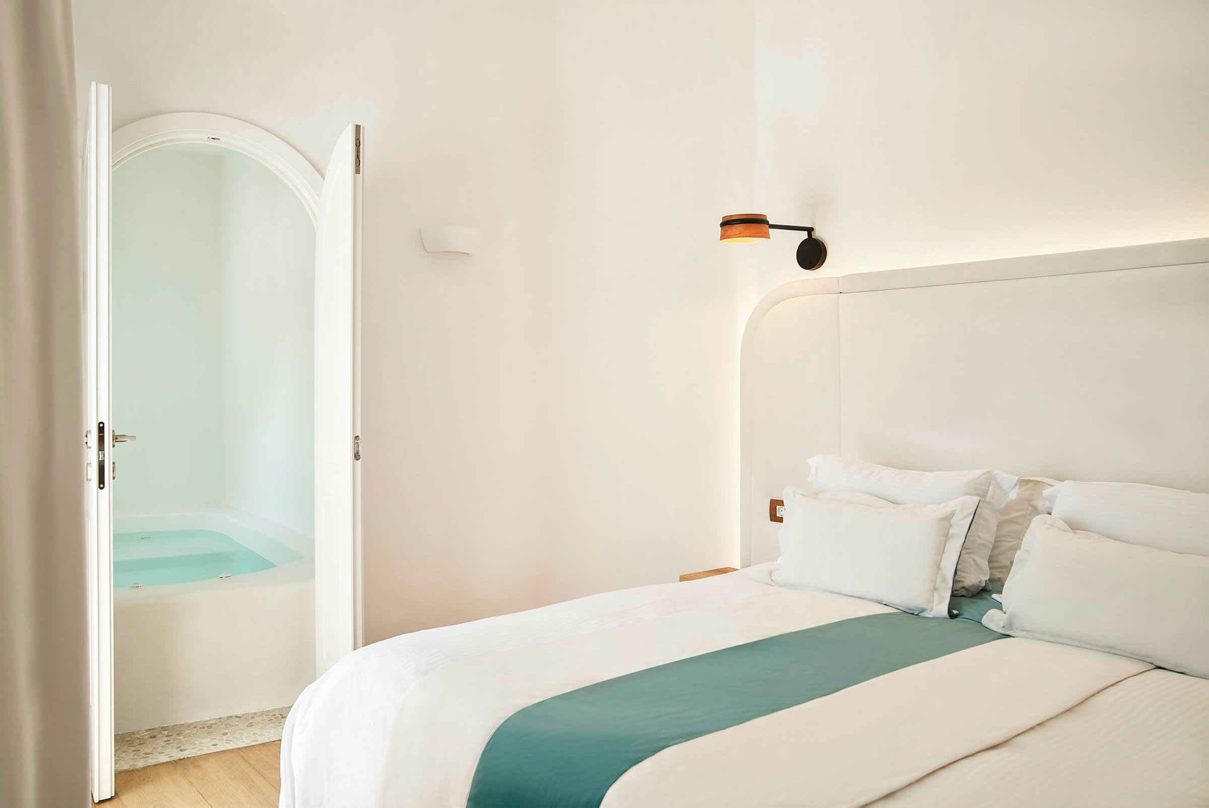 Ett ljust sovrum med en dubbelsäng och öppna dörrar som leder till ett rum med en jacuzzi. (AI-genererad text)